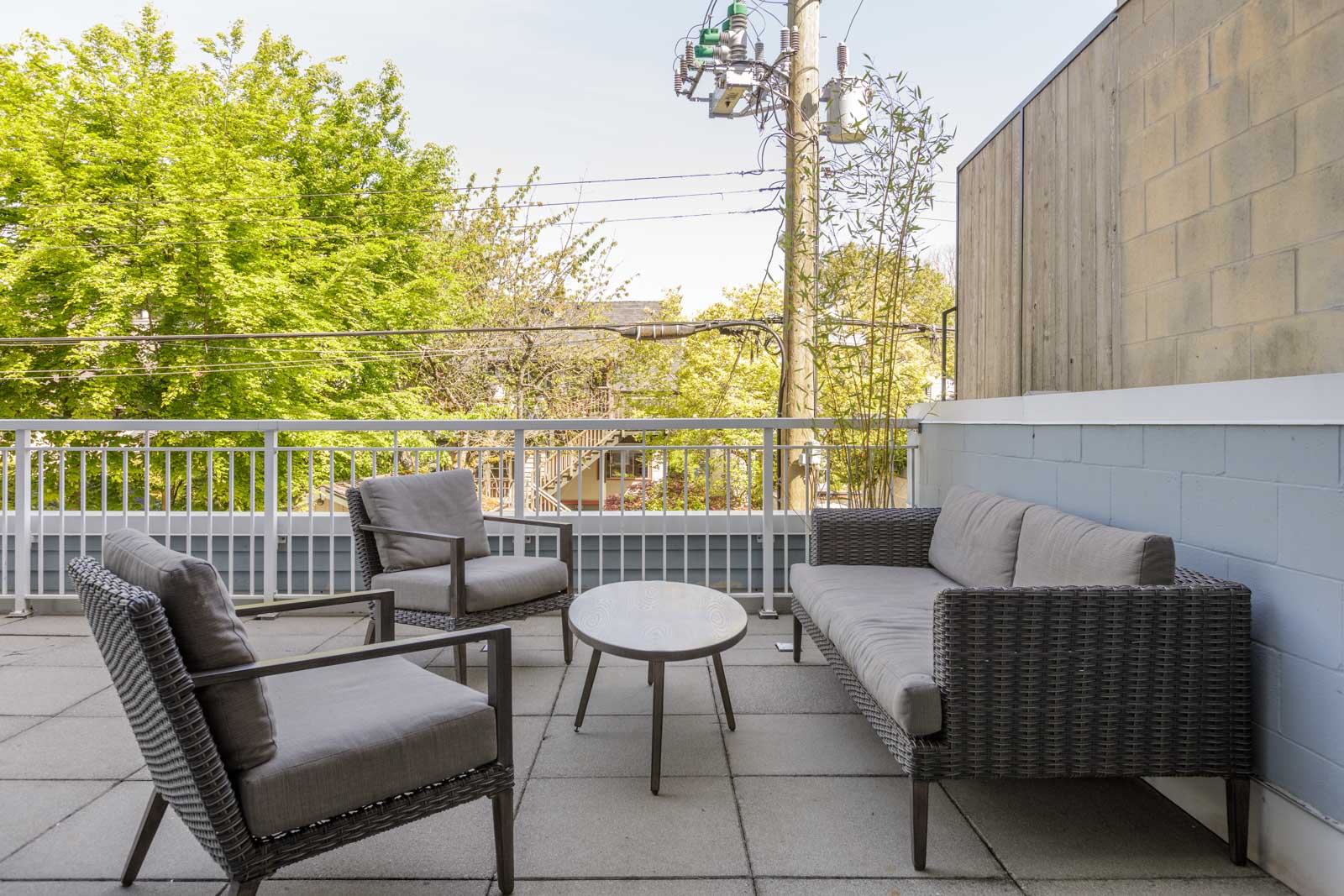Outdoor patio with a wicker sofa, two armchairs, and a round table on a tiled floor, set against a railing and wall, with trees and power lines in the background.