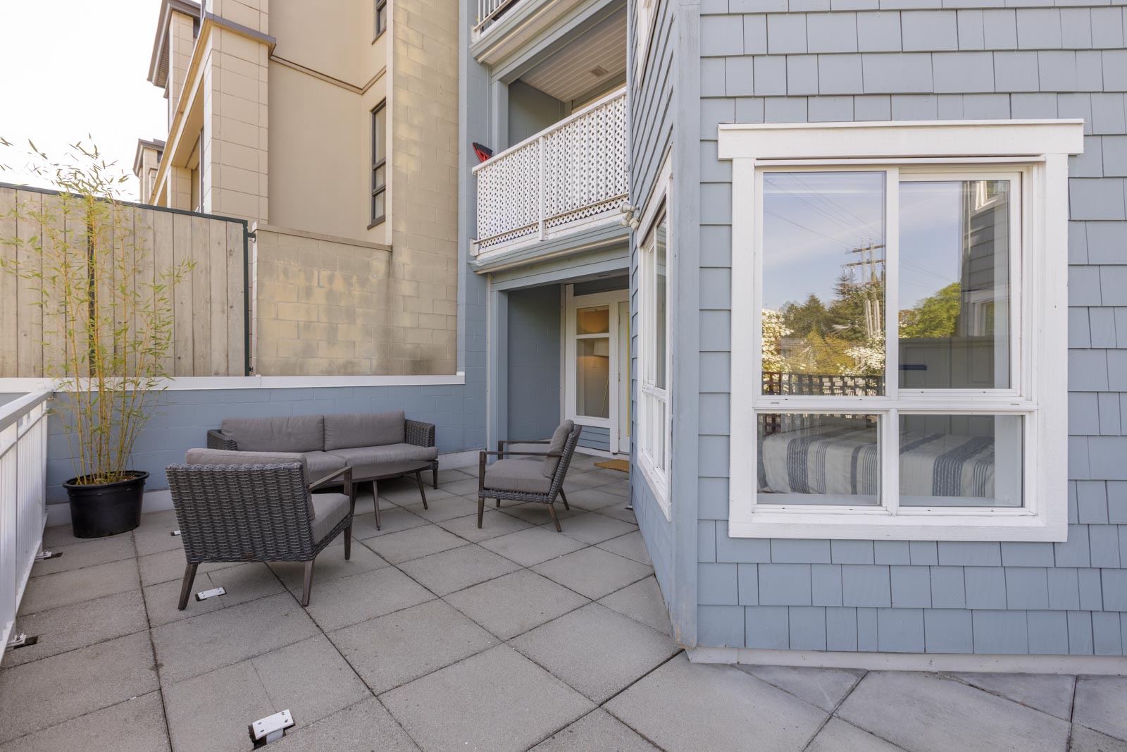A small outdoor patio with gray tile flooring, a potted bamboo plant, a gray sofa, two chairs, and a table next to a blue-sided building with white-trimmed windows.
