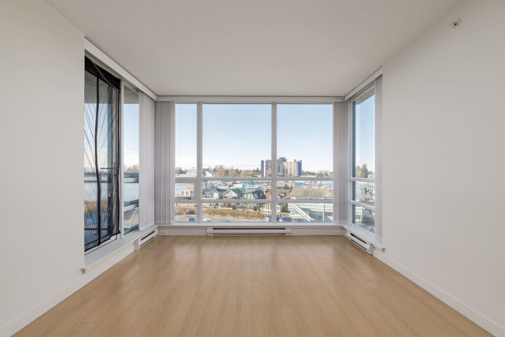 Empty bright room with light wood flooring, white walls, and large floor-to-ceiling windows overlooking a cityscape and distant water.