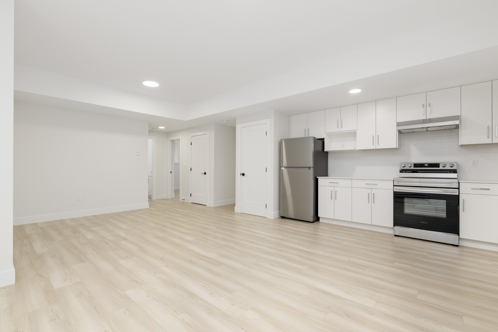 A modern kitchen and living space with light wood flooring, white cupboards, stainless steel appliances, and recessed lighting.