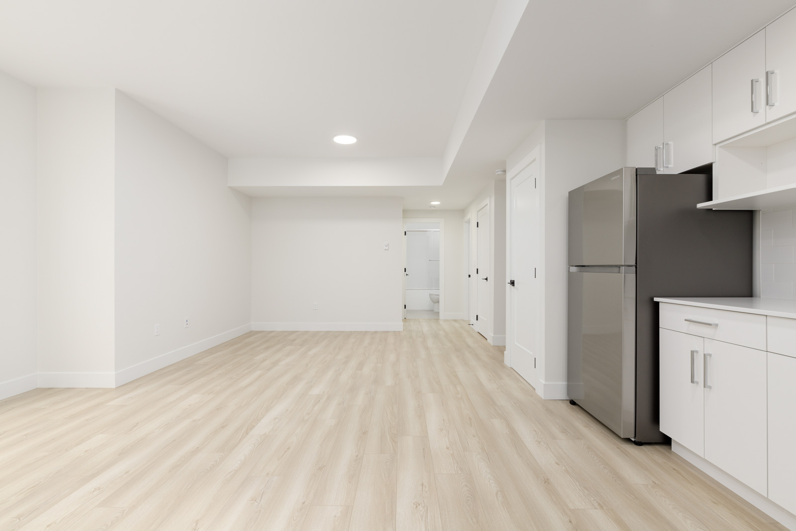 Unfurnished room with light wood flooring, white walls, and a kitchenette featuring white cabinets and a stainless steel refrigerator.