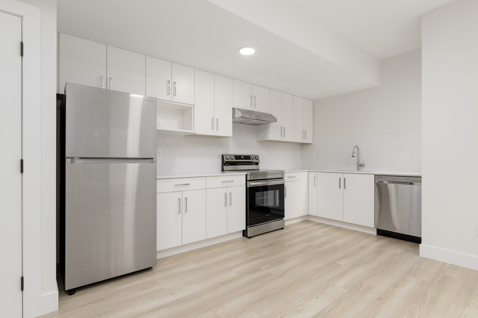 Modern kitchen with white cabinets, stainless steel appliances including a fridge, oven, and dishwasher, and light wood flooring.