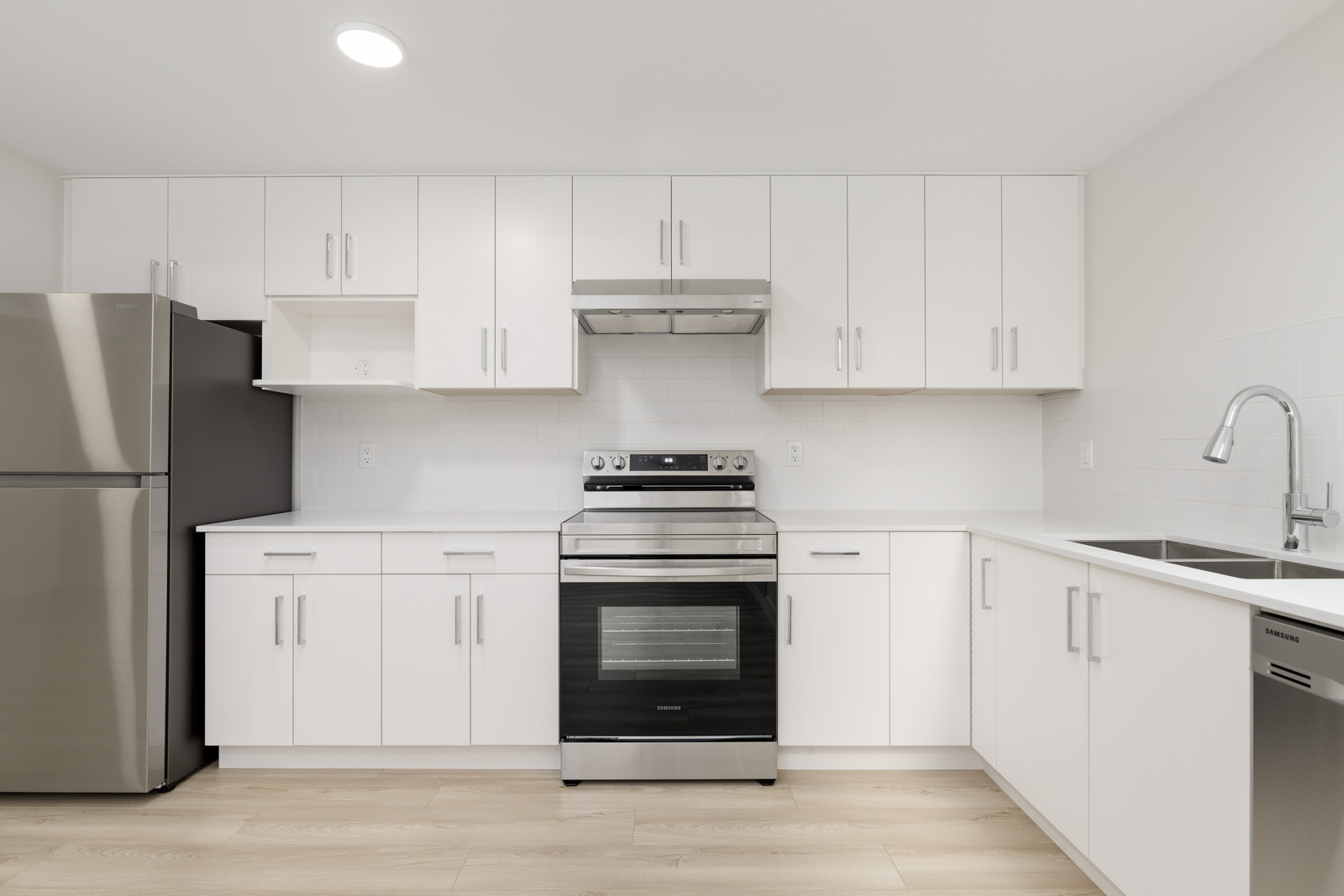 Modern kitchen with white cabinets, stainless steel appliances, and light wood flooring. Includes fridge, oven, range hood, sink, and dishwasher. Minimalist design with overhead lighting.
