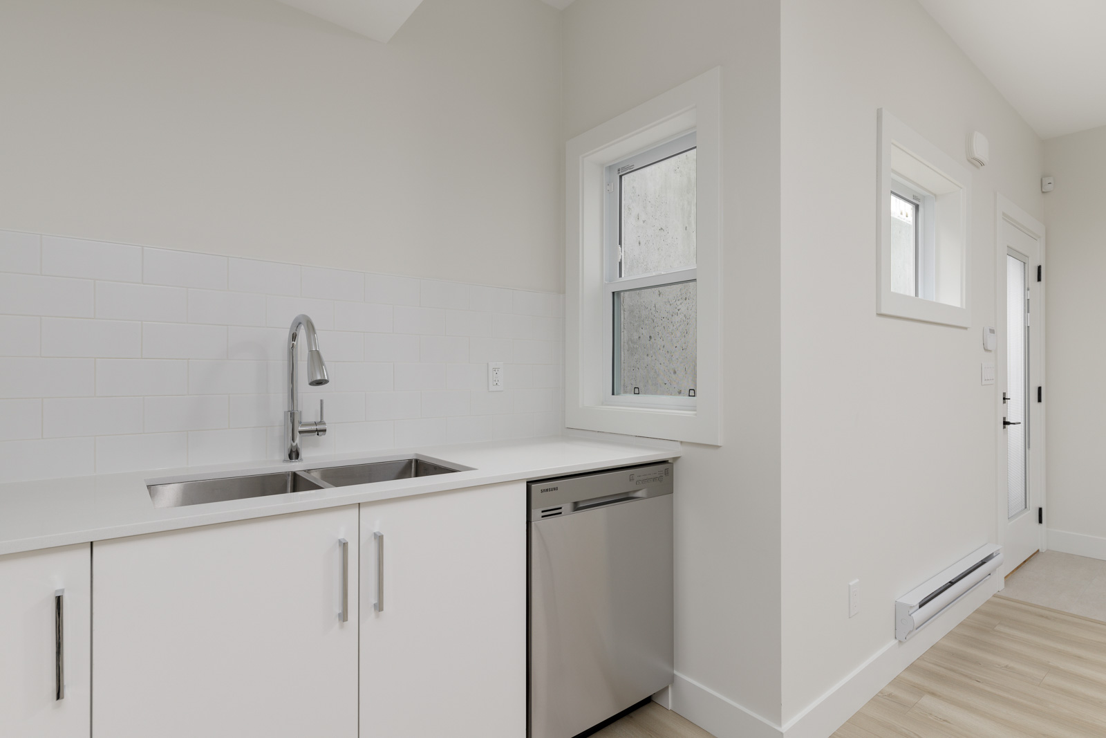 A modern kitchen with white cabinets, a double sink, a stainless steel dishwasher, and a white tiled backsplash. A window and door are visible on the right side of the image.