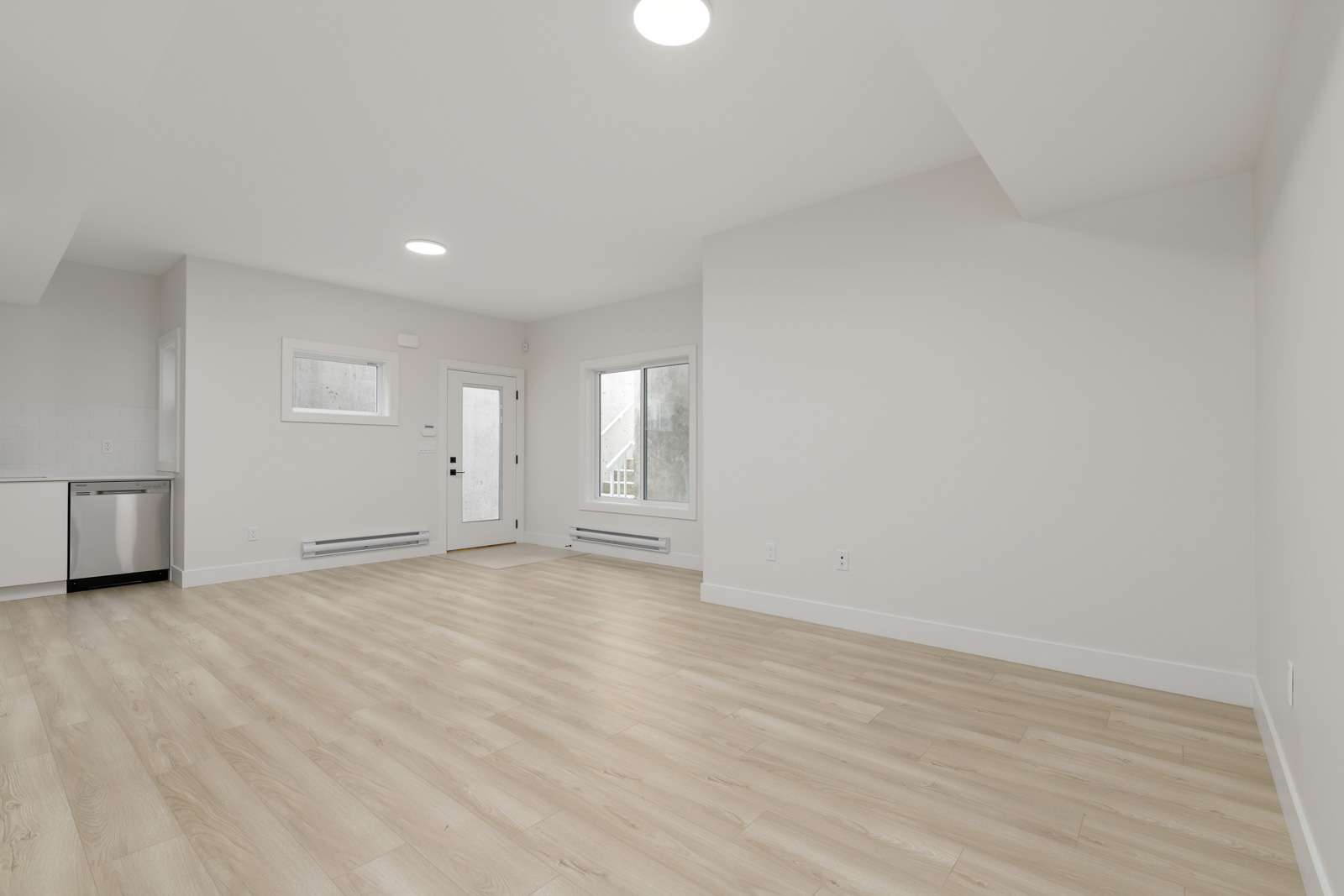 Empty room with light wood flooring, white walls, and a large window. The room includes a visible kitchen area with a dishwasher and a door leading outside.