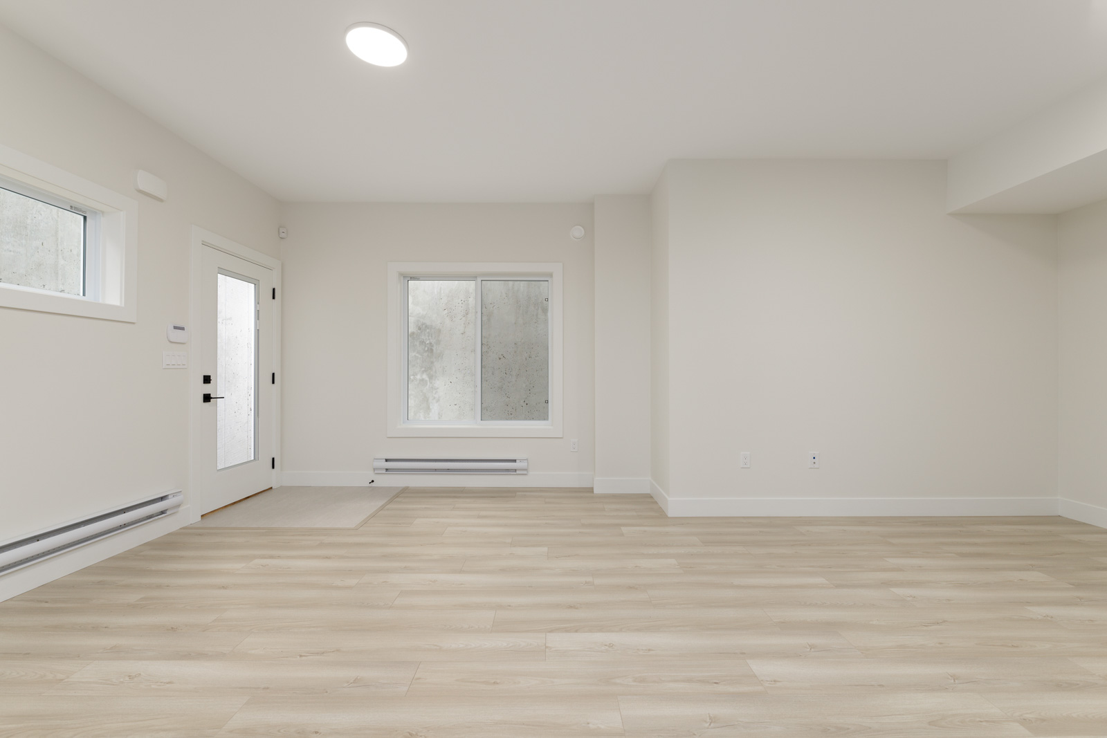 An empty room with light wood flooring, white walls, a window, and a door. Ceiling light and baseboard heater are visible.