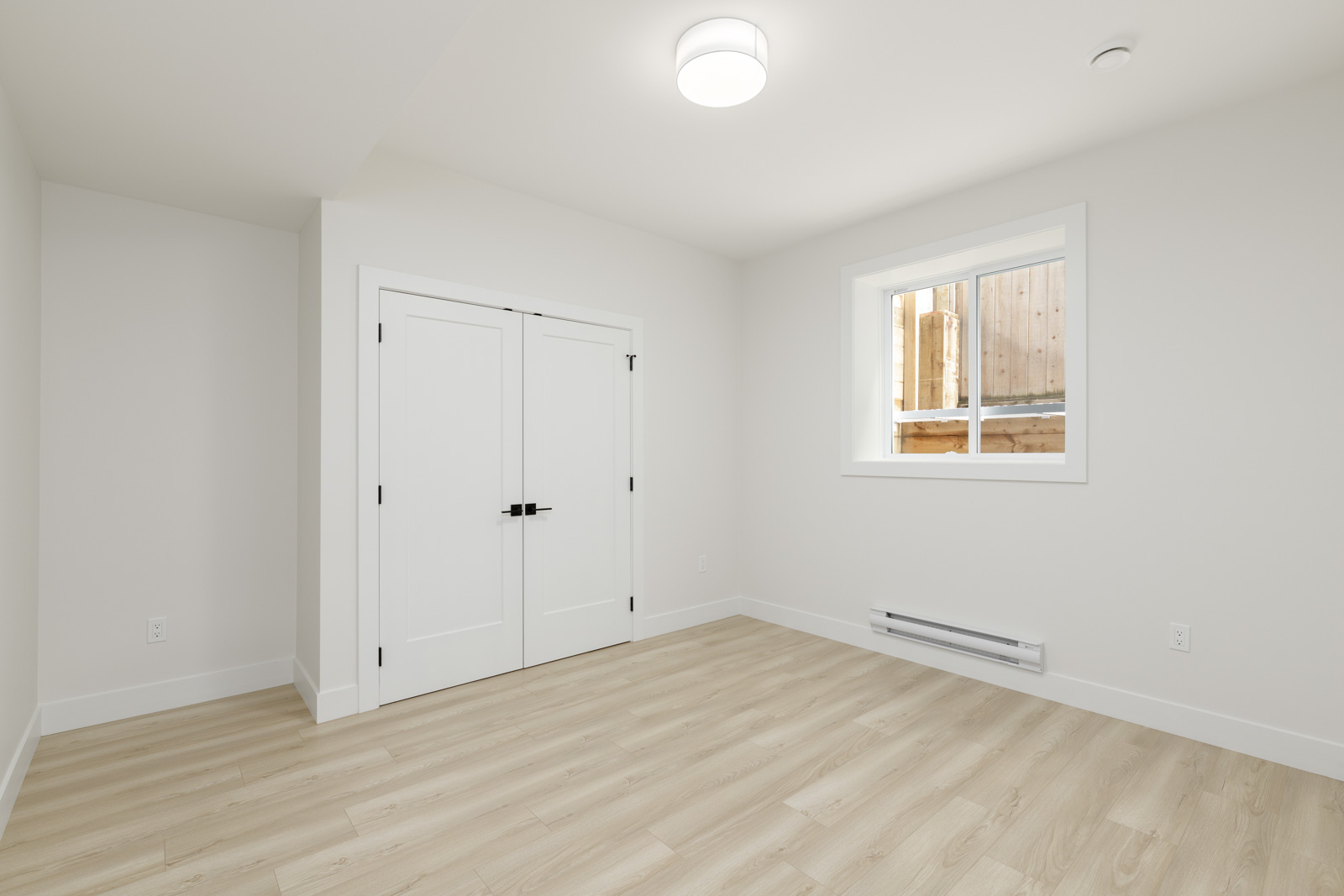 A minimalist white room with wooden flooring, a window, and a closed double door. A modern ceiling light fixture is centered on the ceiling.
