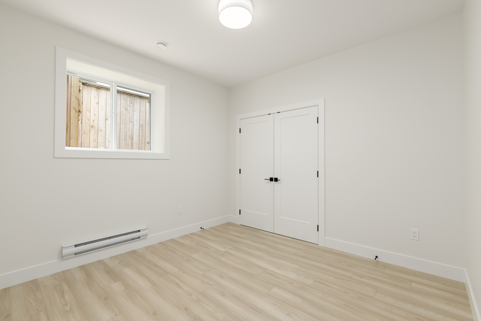 Empty white room with light wooden flooring, double doors, and a small window with a view of a wooden fence. Ceiling light fixture is centered above.
