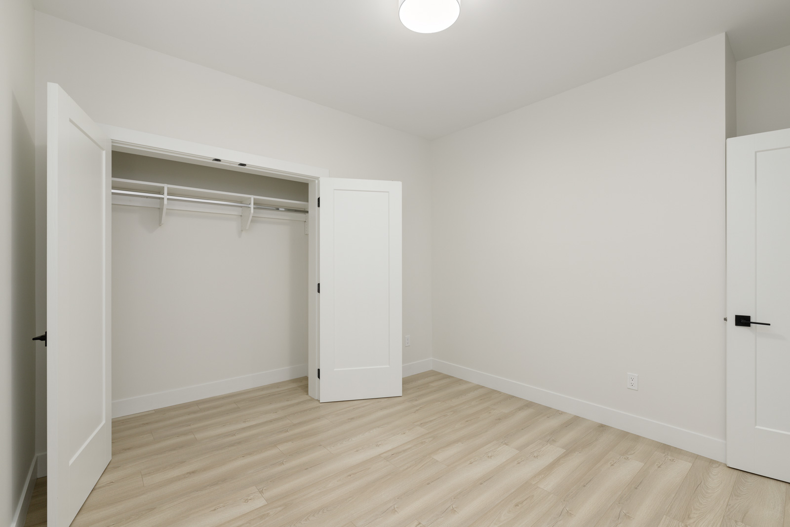 Empty white room with light wood flooring, open closet doors revealing a closet with a hanging rod and shelf. Ceiling light fixture and plain walls are visible.