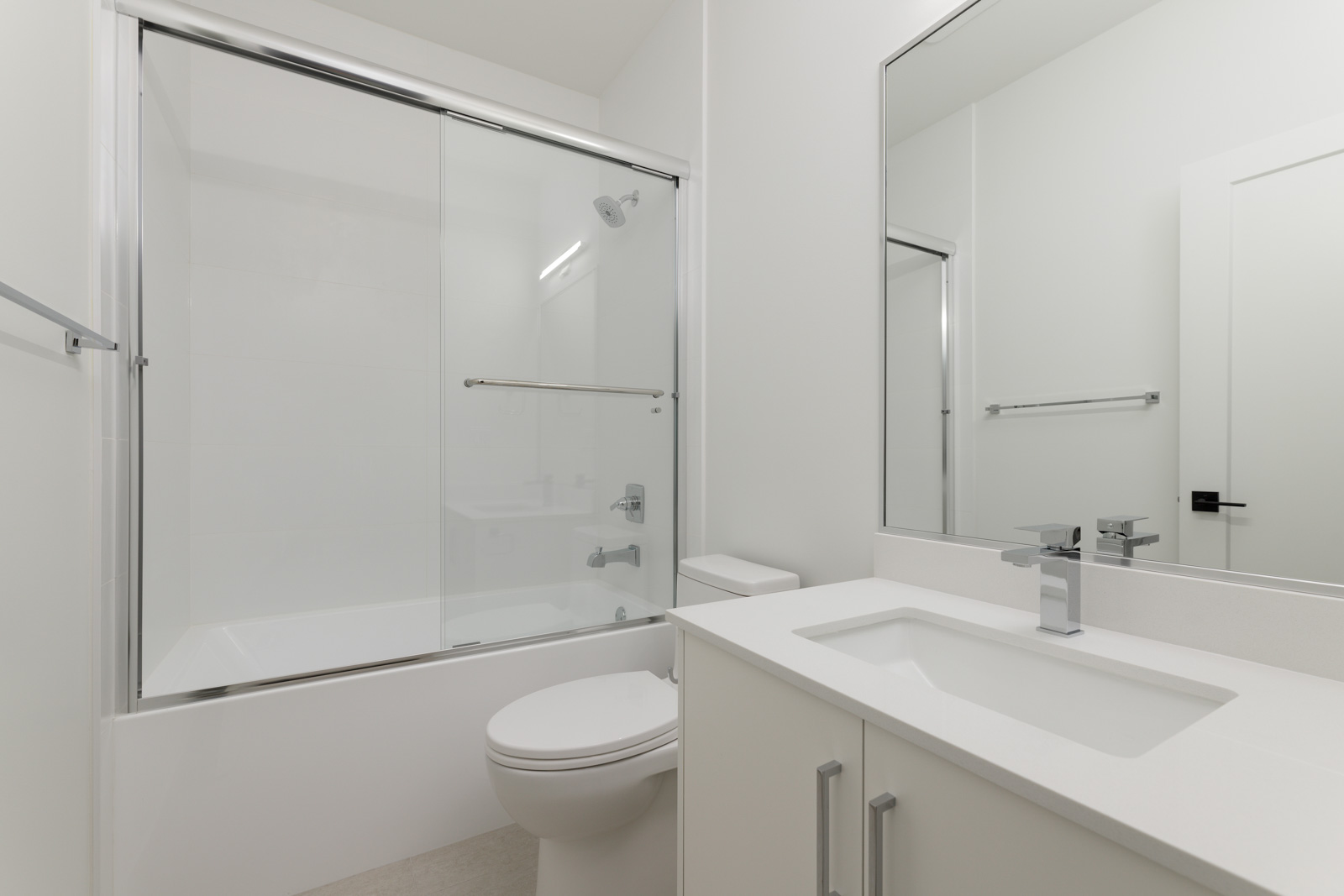 Modern bathroom with a white bathtub and glass sliding door, toilet, and sink with a large mirror above. The room features a minimalist design with white walls and fixtures.