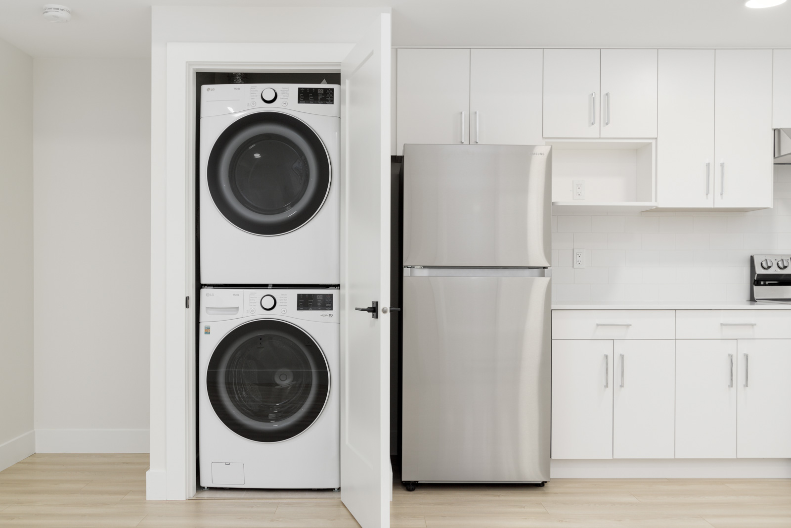 A modern laundry space with a stacked washer and dryer behind a door. Next to it is a stainless steel refrigerator surrounded by white cabinets and countertops.