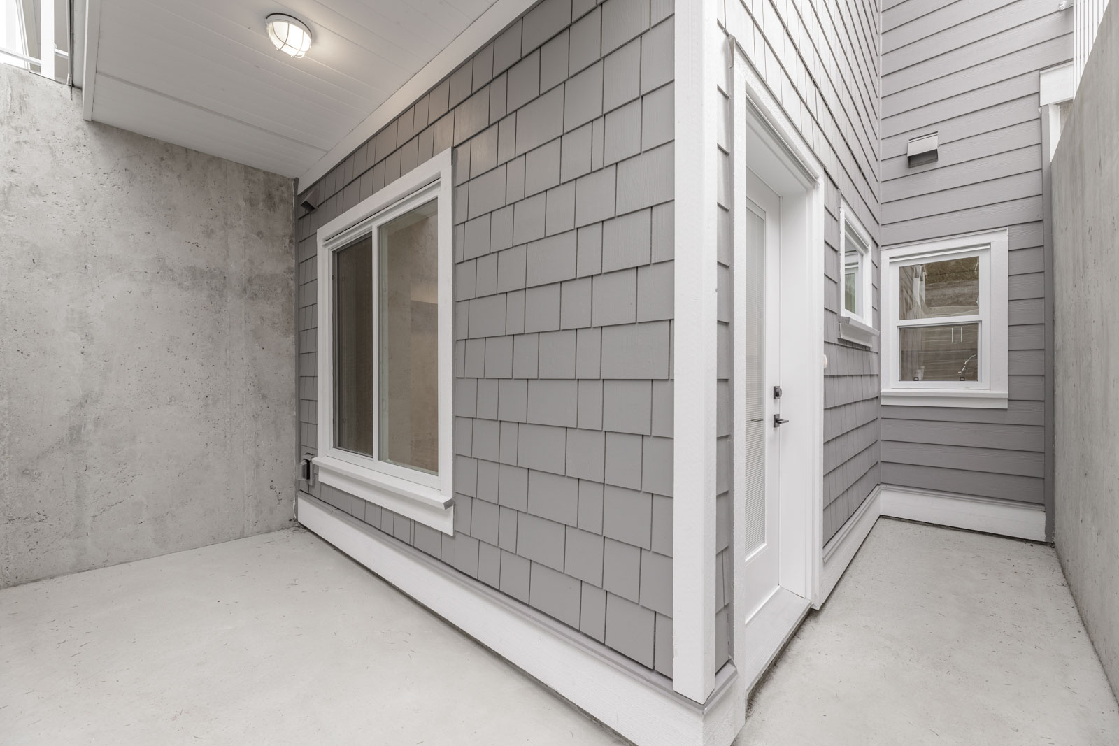 Small, enclosed outdoor area with gray shingle siding, a window, and a white door. Concrete walls and floor, with a light fixture on the ceiling.