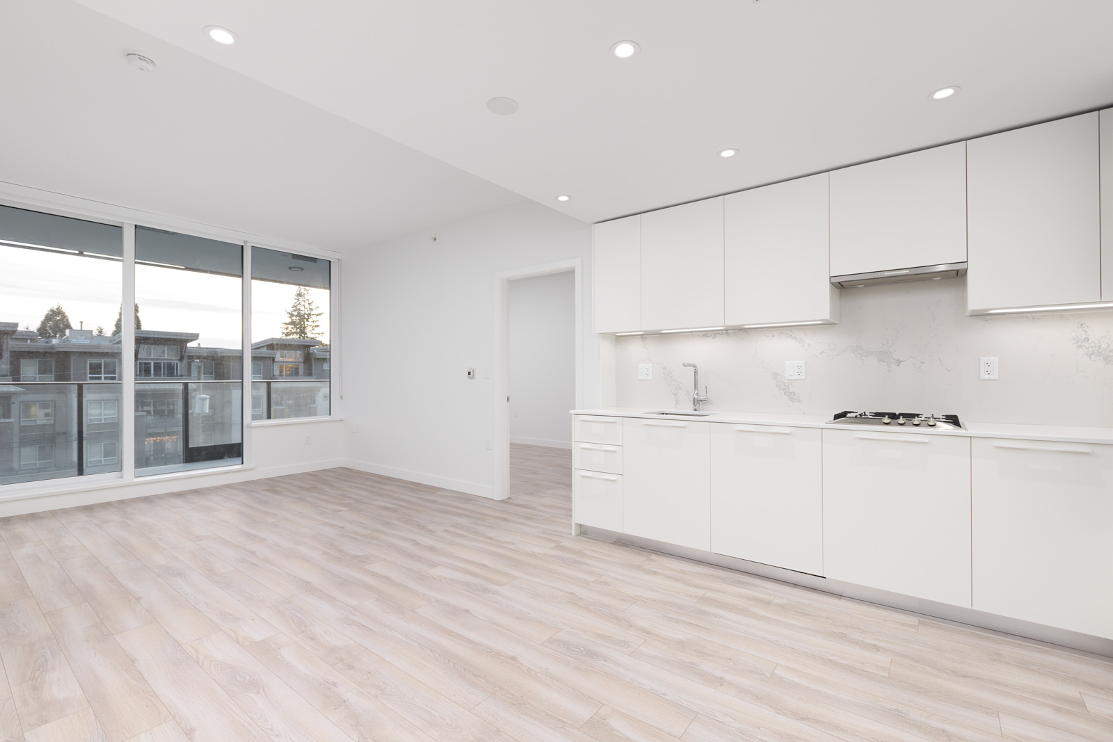 Modern, unfurnished kitchen and living area with light wood floors, white cabinets, and large windows.