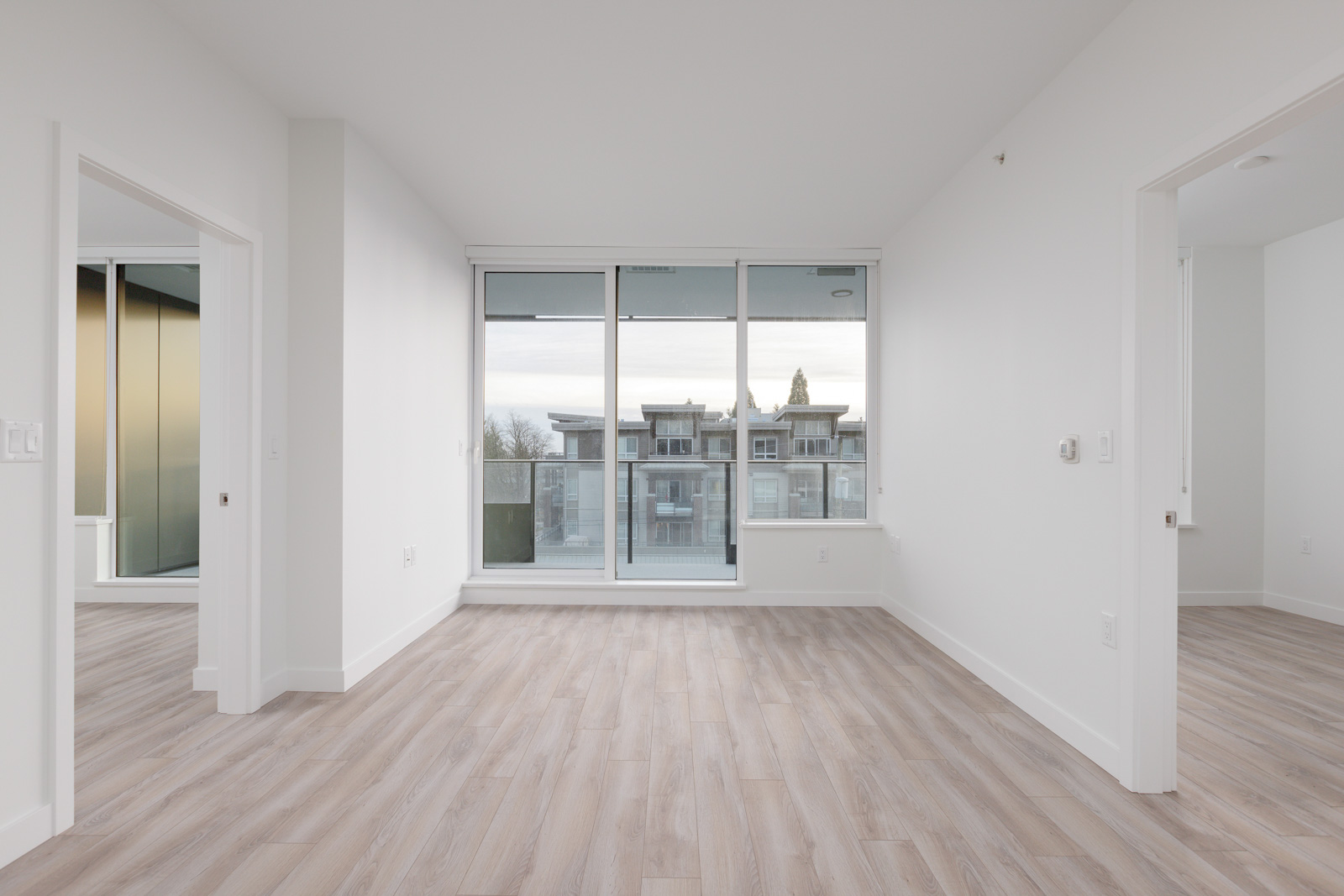 Empty room with light wood flooring and white walls, featuring large glass doors leading to a balcony. Two open doorways on either side of the room.