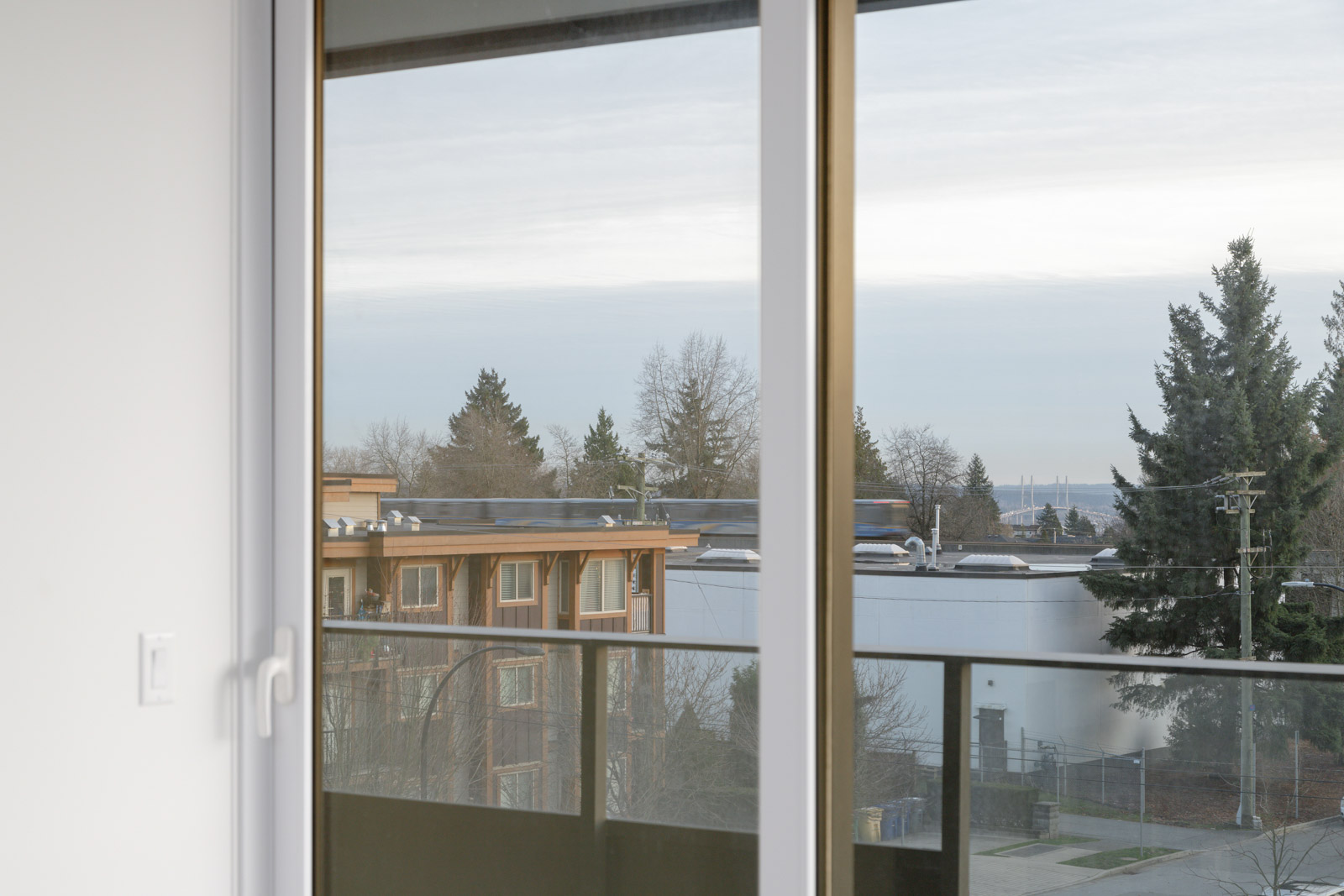 View from a window showing a balcony, distant trees, a residential building, and a partly cloudy sky.