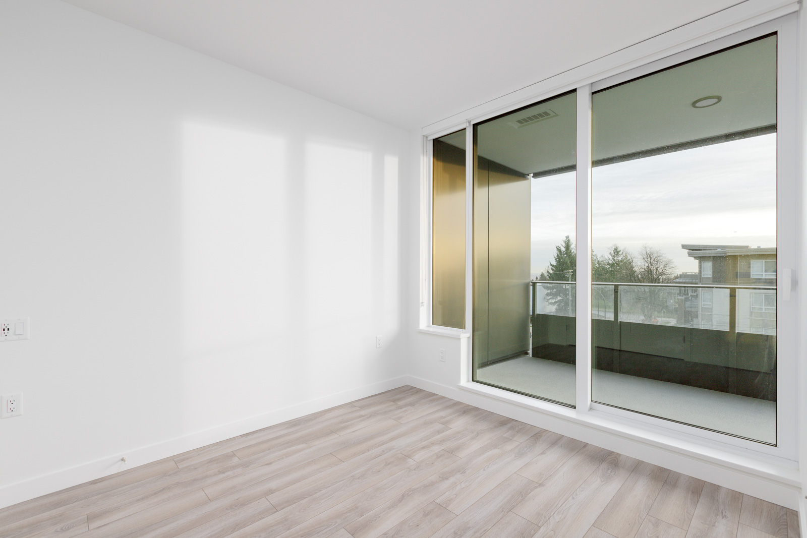 Empty room with light wood flooring, white walls, and large sliding glass doors leading to a balcony. Natural light enters through the doors, casting shadows on the wall.