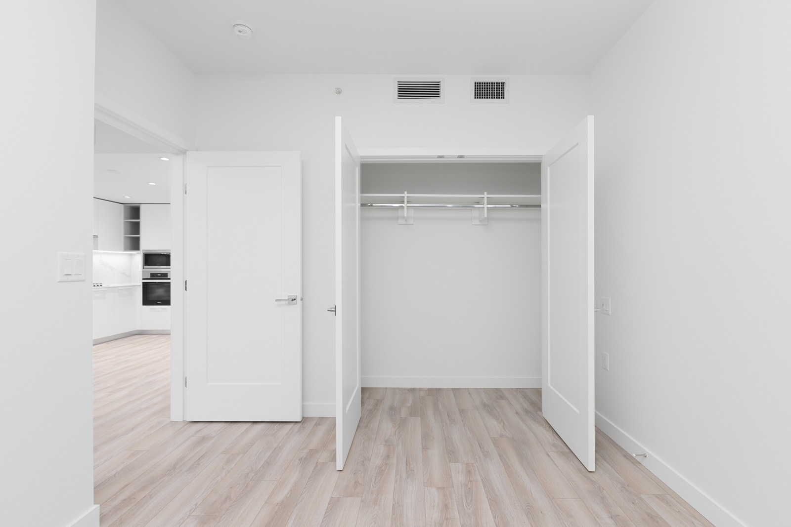 Empty room with open closet doors, light wood flooring, and a view into a modern kitchen with white cabinets and appliances.