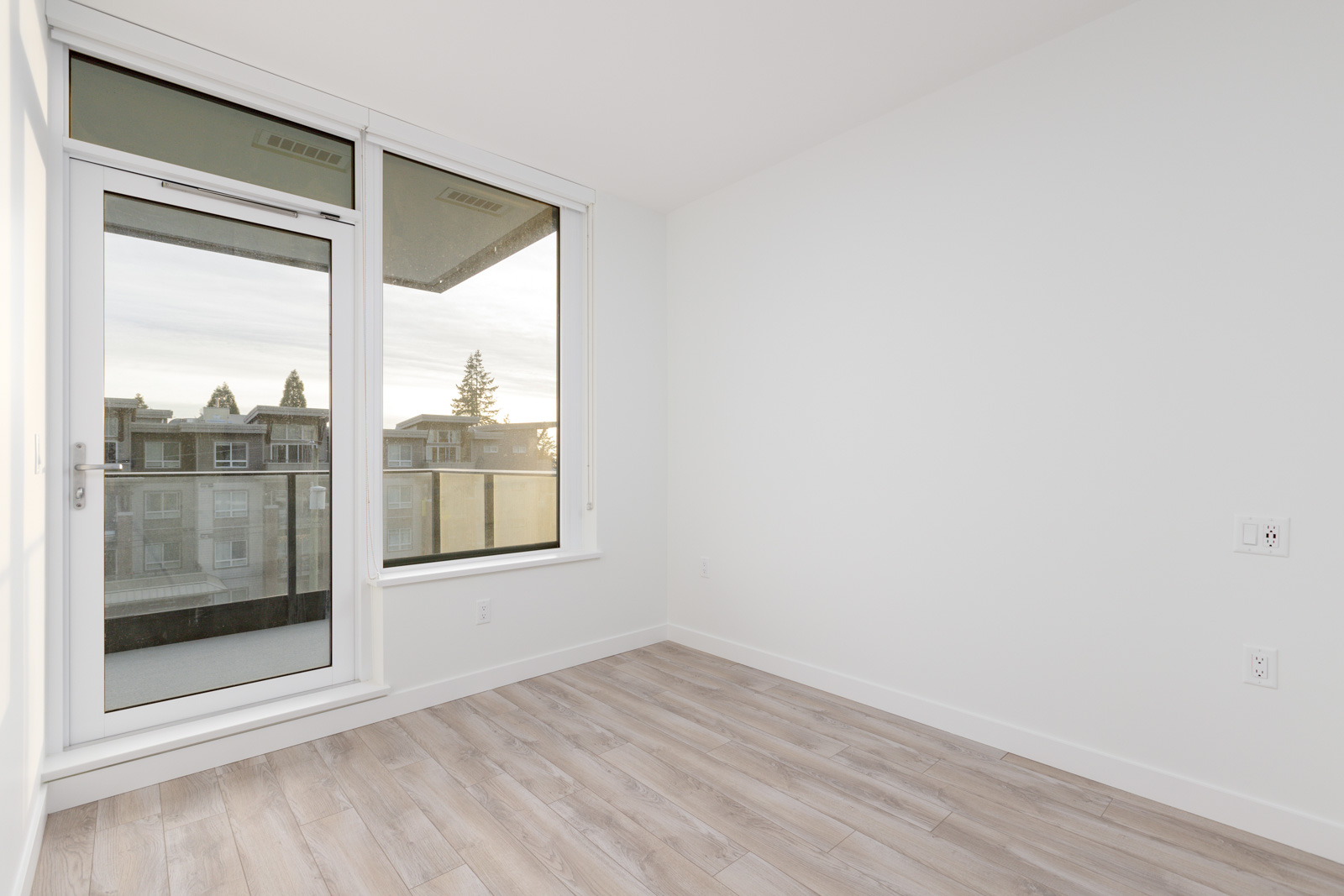 Empty room with light wood flooring, large window, and glass door leading to a balcony. Walls are white, and natural light illuminates the space.