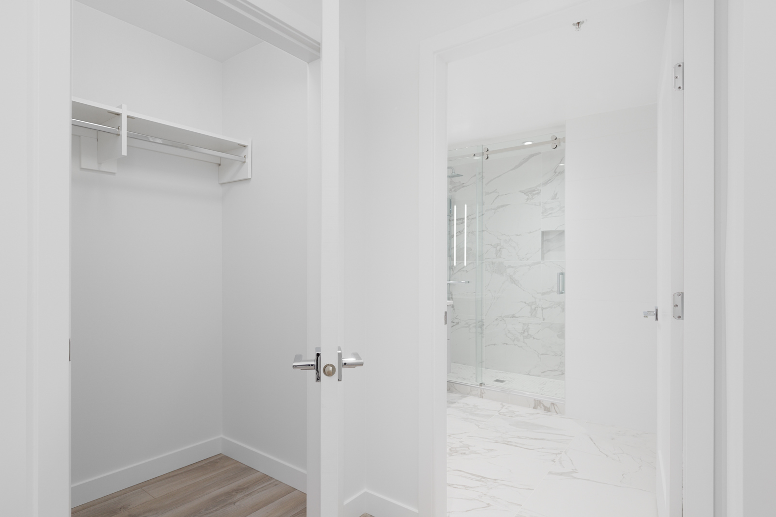 Open door reveals an empty closet with a wooden floor on the left and a bright bathroom with marble walls and a glass shower door on the right.