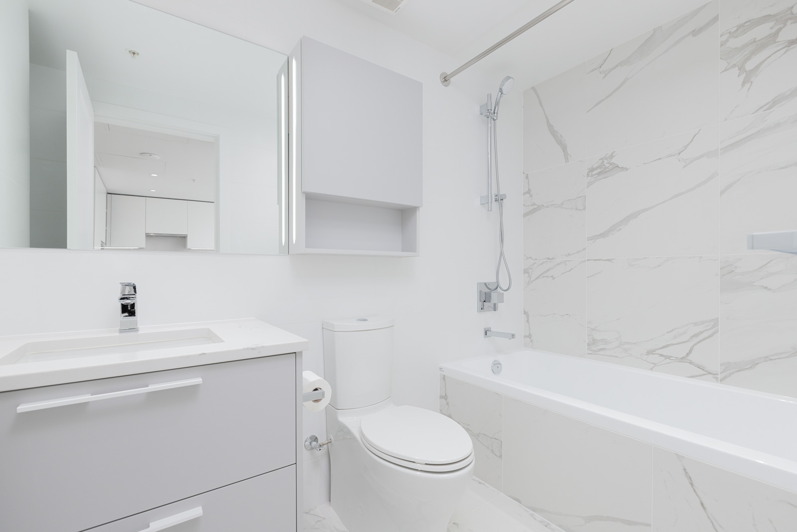 Modern white bathroom with a bathtub, shower, toilet, and sink. Large mirror above the sink and marble-patterned tiles on the walls.
