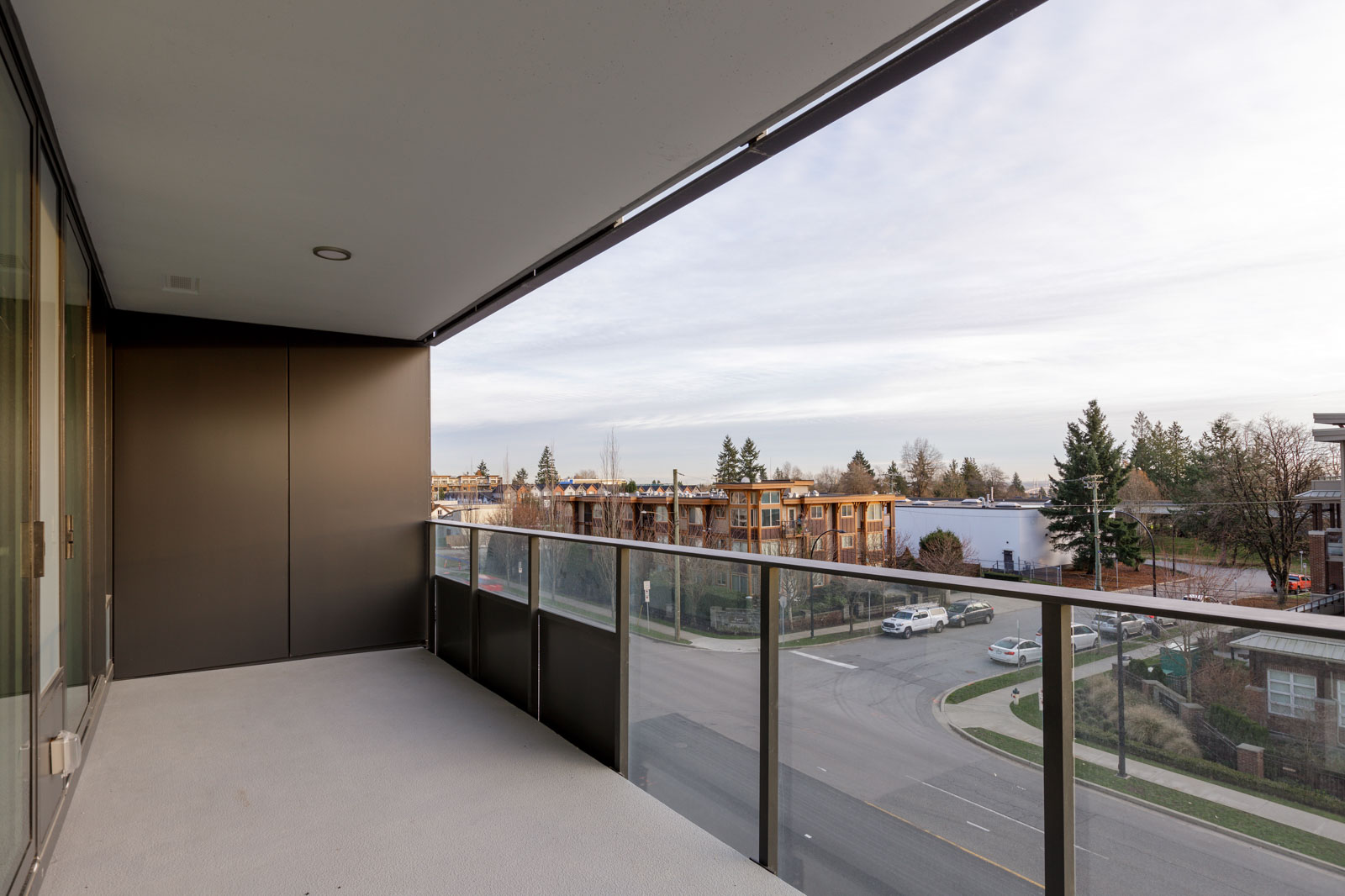 A modern balcony with glass railing overlooks a quiet suburban street and residential buildings under a cloudy sky.