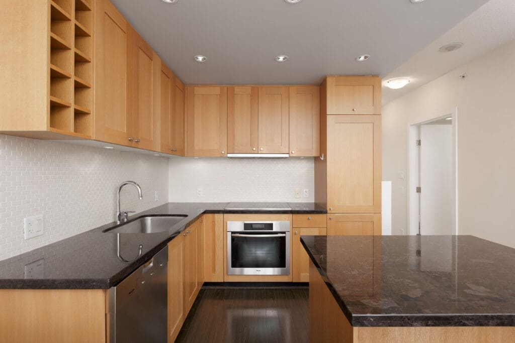 Modern kitchen with light wood cabinets, dark countertops, stainless steel oven, built-in sink, and a kitchen island under recessed lighting.