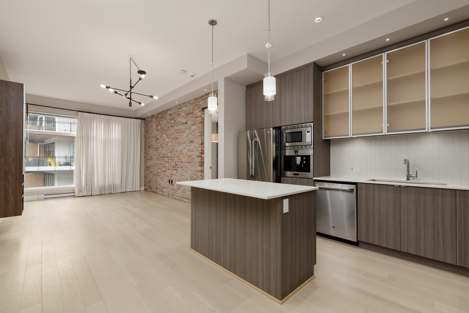 Modern kitchen with a central island, wooden cabinets, stainless steel appliances, and a brick accent wall. Large window with curtains on the left side.