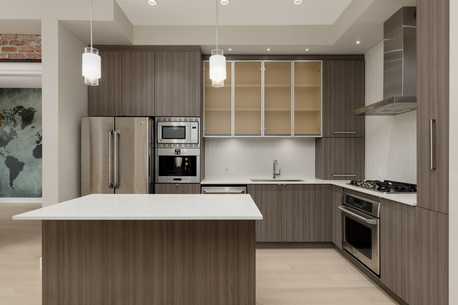 Modern kitchen with wooden cabinets, stainless steel appliances, and a white island countertop. Hanging pendant lights and a partial world map mural are visible.