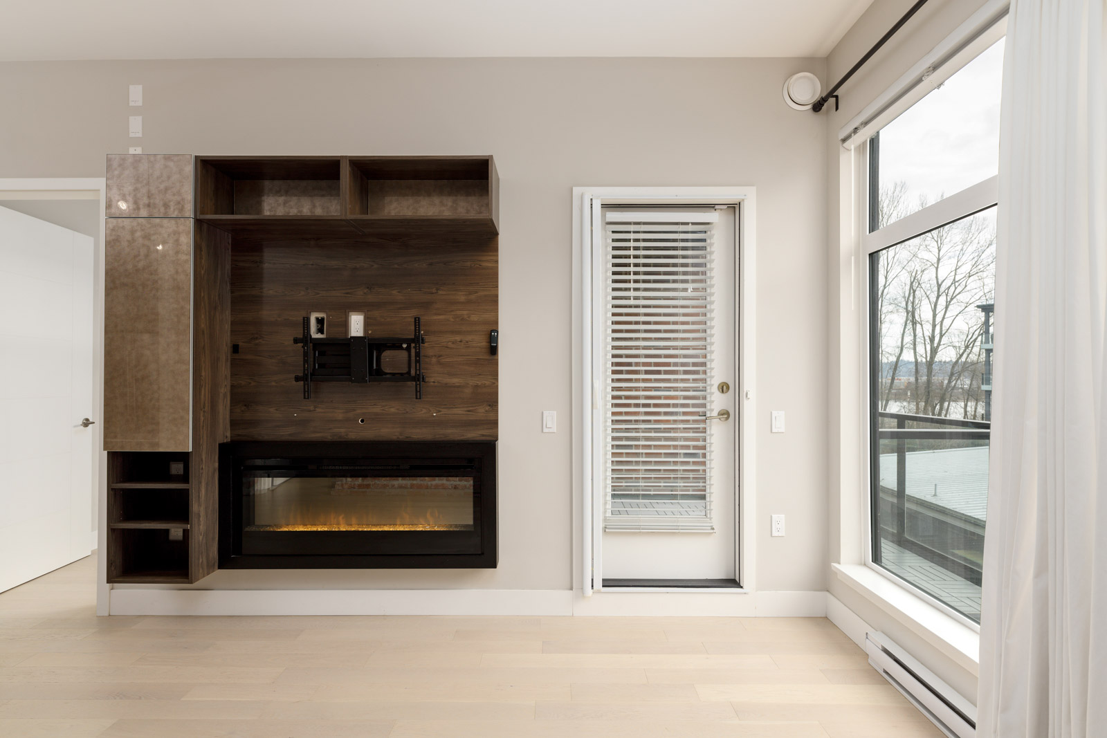 Modern living room with a mounted TV space, fireplace, and shelving on the left wall; a glass door with blinds leads to an outdoor view on the right. Light wood flooring and neutral walls.