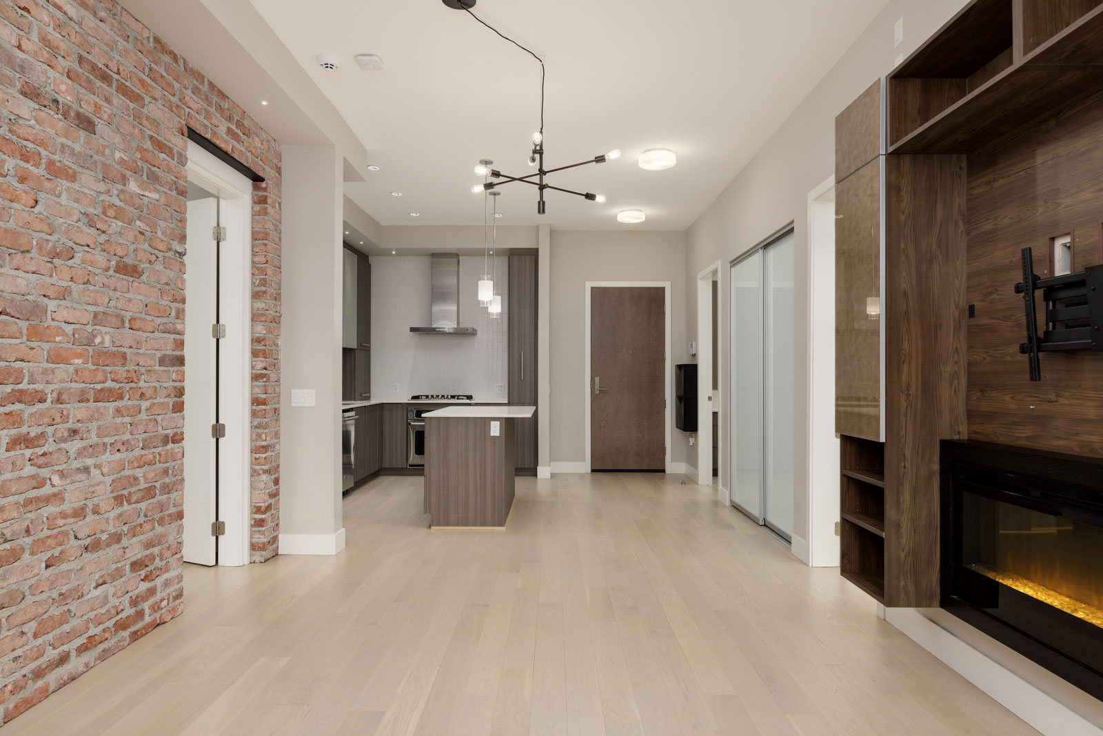 Modern living area with light wood floors, an exposed brick wall, and a contemporary kitchen. Features built-in wooden shelves, a fireplace, and a minimalistic chandelier.