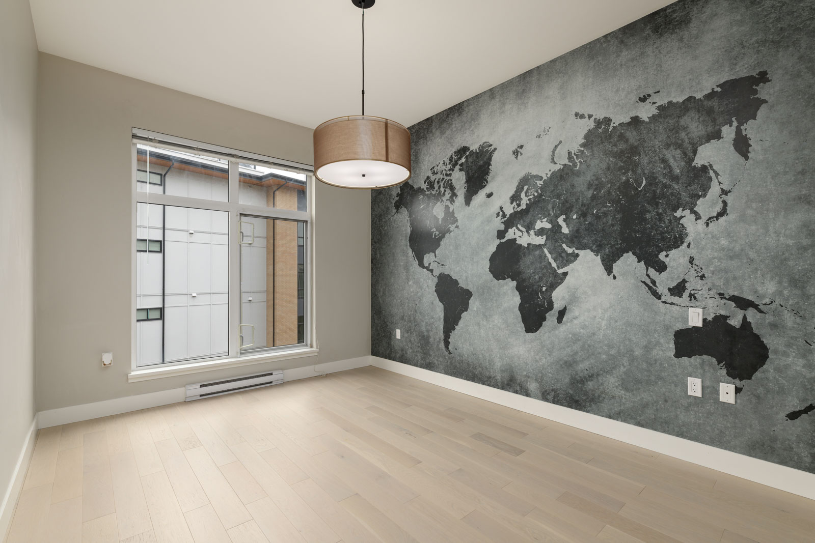 Empty room with light wooden flooring, a window on the left, a ceiling light, and a wall featuring a black world map mural.
