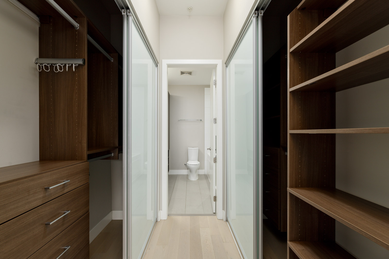 Walk-in closet with wooden shelves and drawers on both sides leading to a bathroom with a view of a toilet.