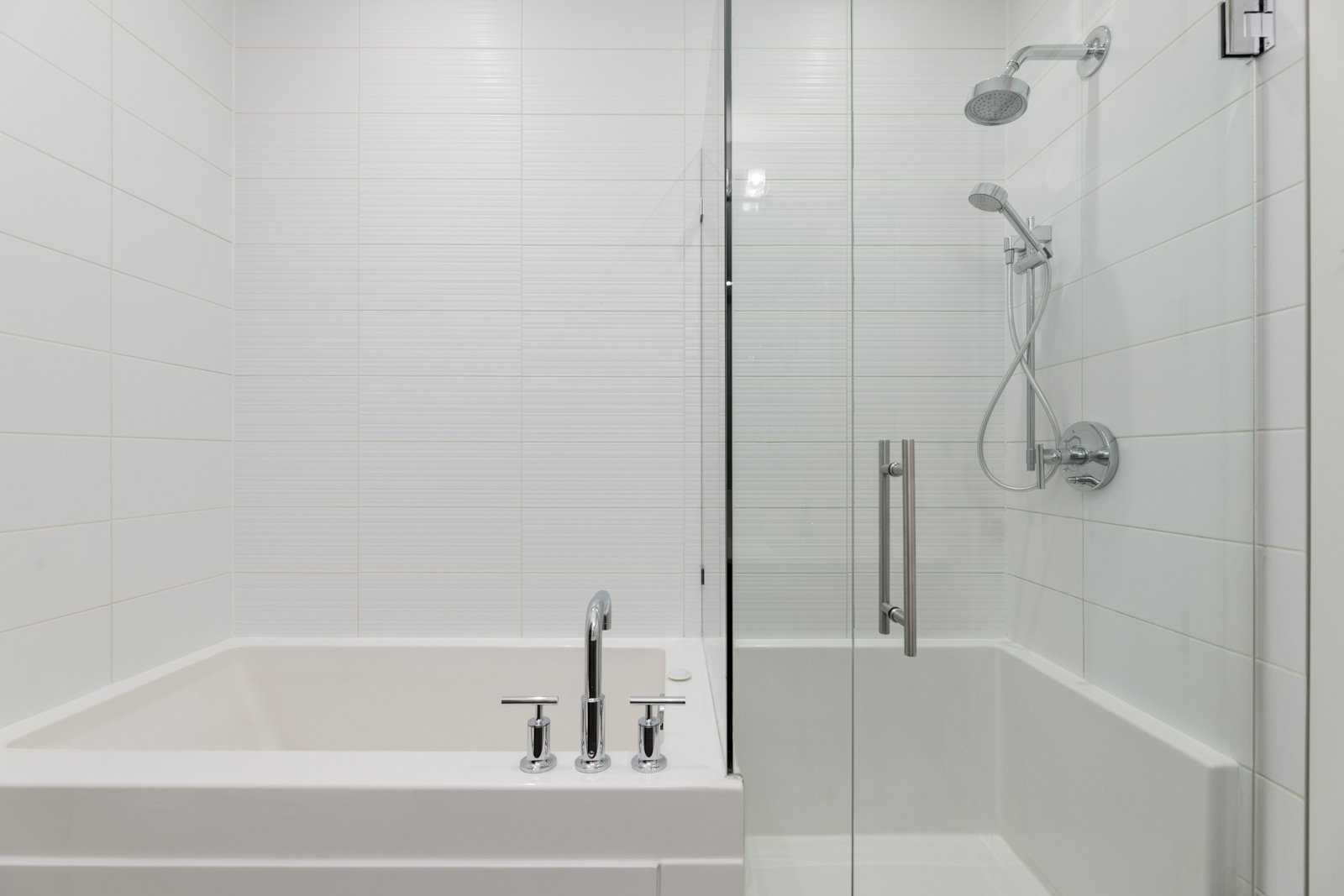 A modern bathroom with a white bathtub and a glass-enclosed shower featuring a rainfall showerhead and wall-mounted controls. The walls are tiled in white.