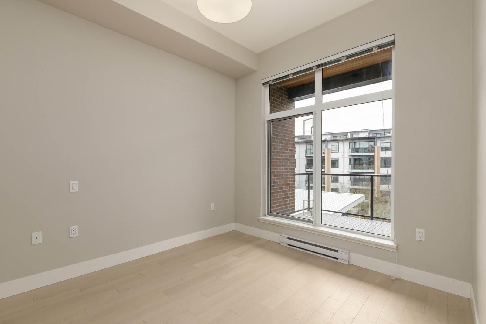 Empty room with beige walls and wooden floor, featuring a large window with a view of apartment buildings. A ceiling light fixture is also visible.