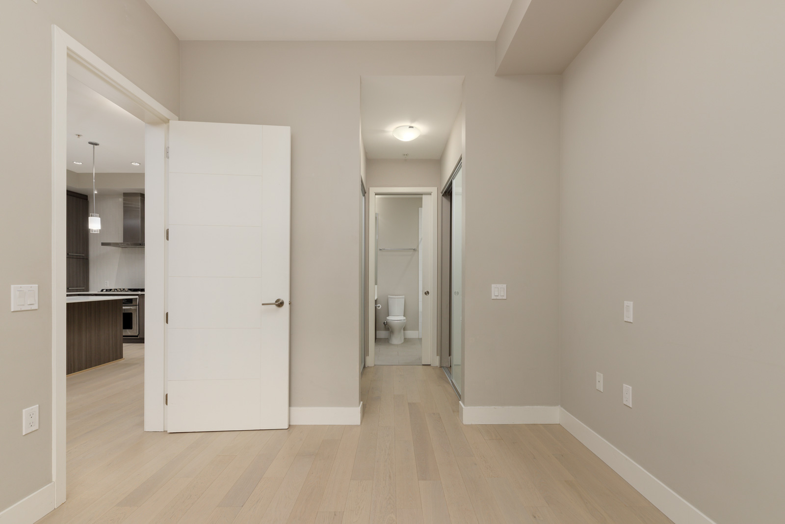 A vacant room with beige walls and light wood flooring, showing an open door to a kitchen on the left and a doorway leading to a bathroom with visible toilet straight ahead.