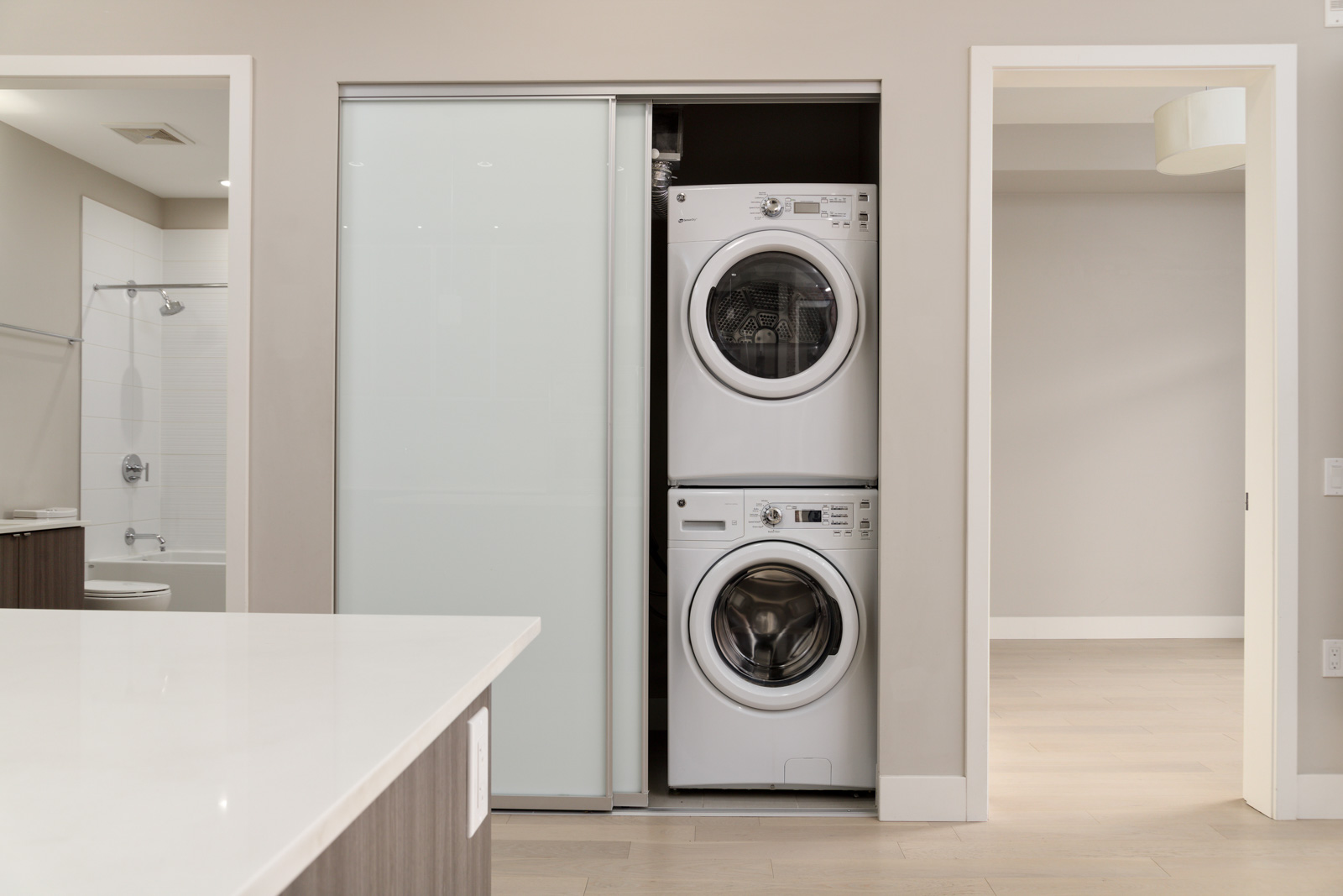 A modern laundry area with stacked washer and dryer units is partially concealed by a sliding white door. Nearby, a light gray bathroom is visible.
