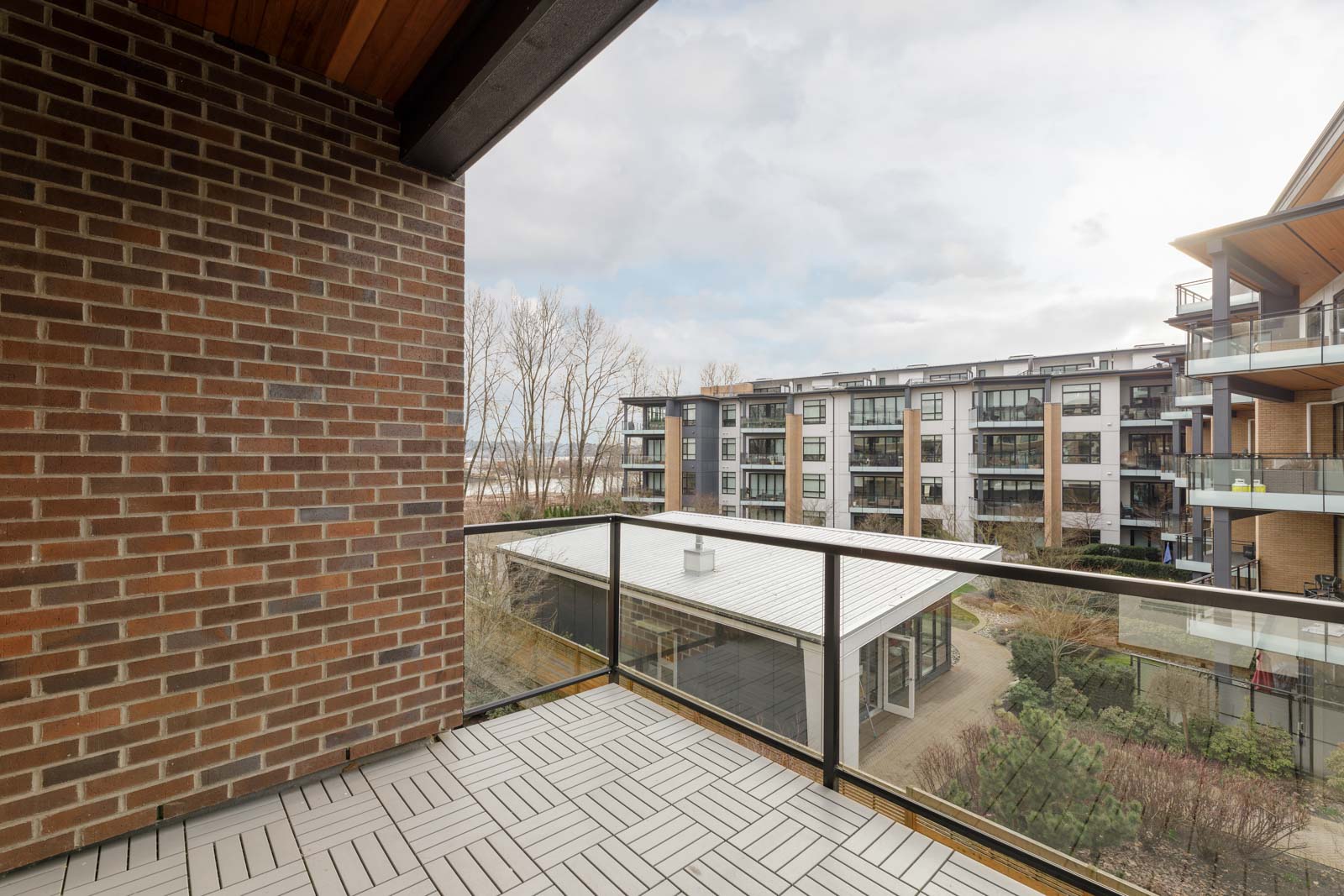 View from a balcony with a brick wall, overlooking a courtyard, trees, and a modern apartment building under a cloudy sky.