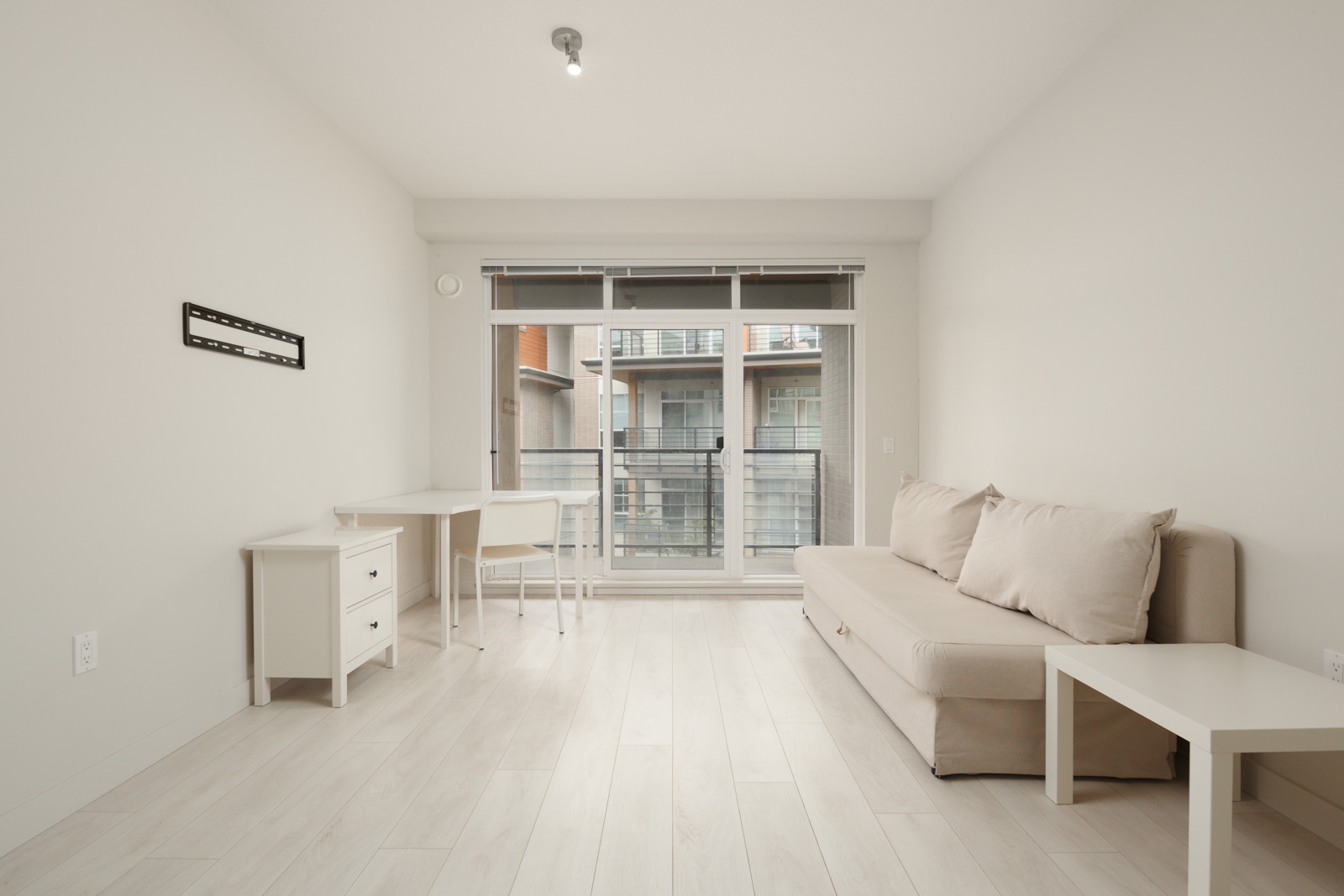 Minimalist room with light wood flooring, featuring a small desk with a chair, a wall-mounted frame, and a beige sofa. Large windows let in natural light, overlooking adjacent buildings.