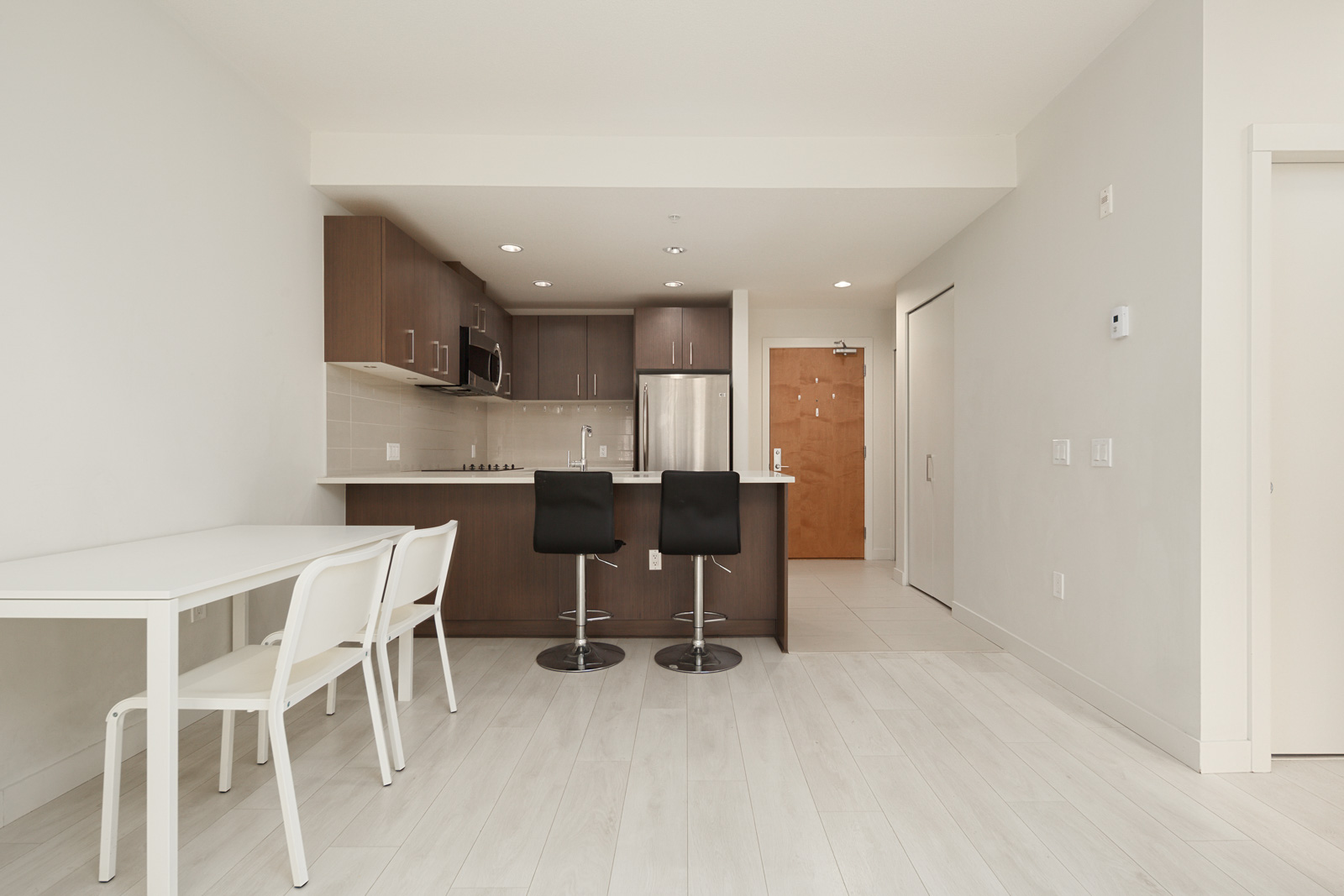 Modern kitchen and dining area with wood cabinets, bar stools at a counter, white dining table with chairs, light wood flooring, and an open doorway.