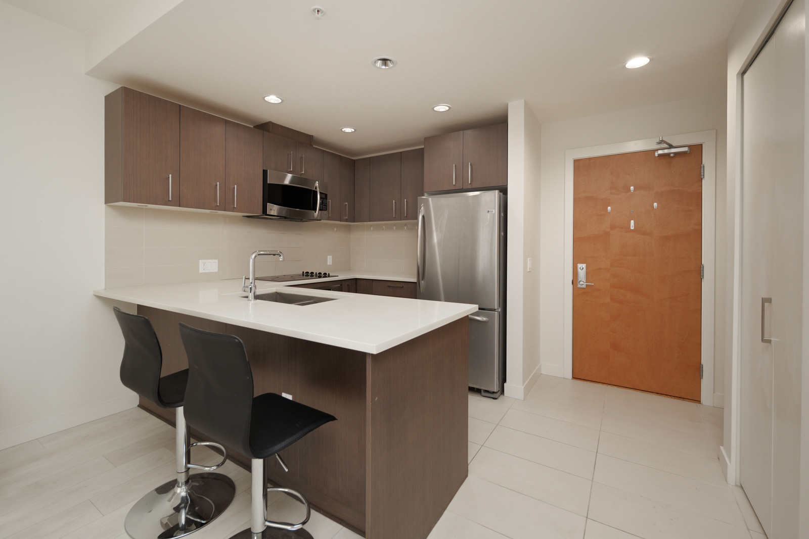Modern kitchen with dark wood cabinets, stainless steel appliances, island with white countertop, two black bar stools, and pale tile floor. Brown door and a white closet are visible.