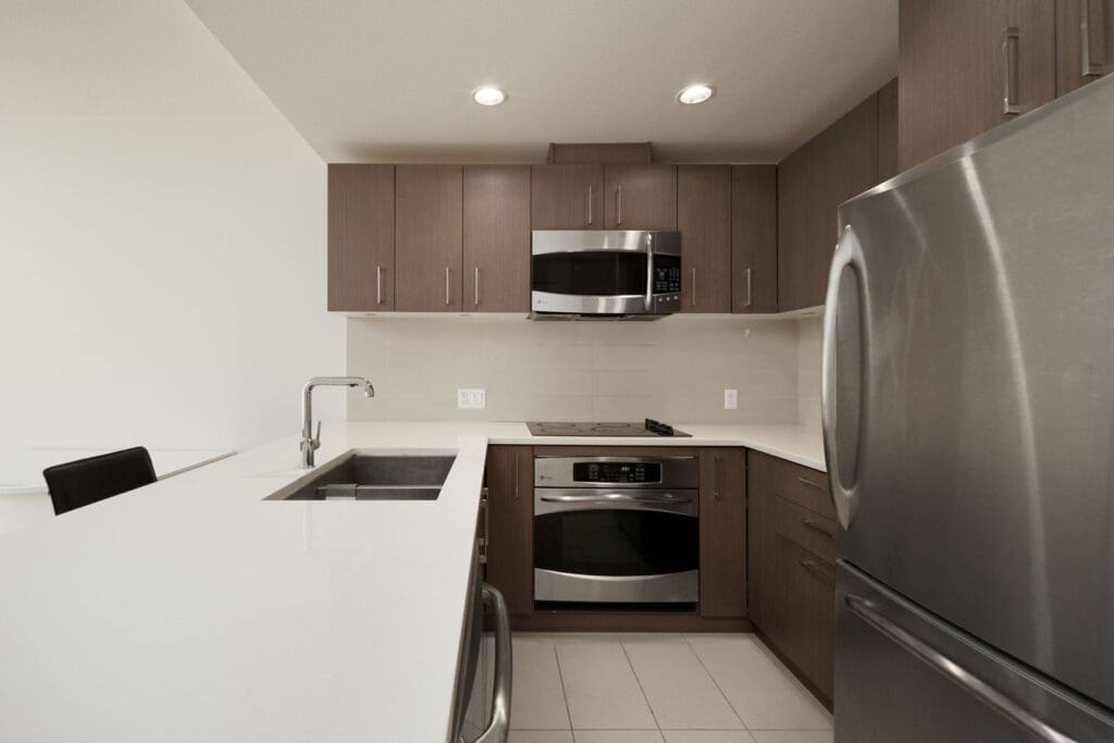 Modern kitchen with dark wood cabinets, stainless steel appliances, white countertops, and tiled flooring. Features built-in oven, microwave, fridge, and sink with faucet.