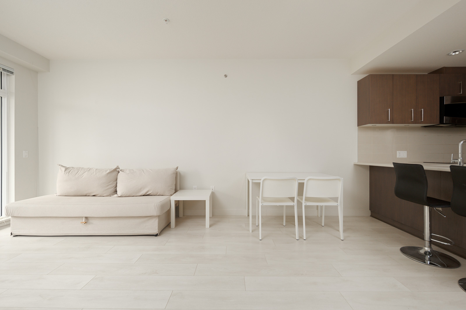Minimalist living area with a beige sofa, white side table, small dining table with two white chairs, and a modern kitchen with bar stools. Walls and floors are light-colored.