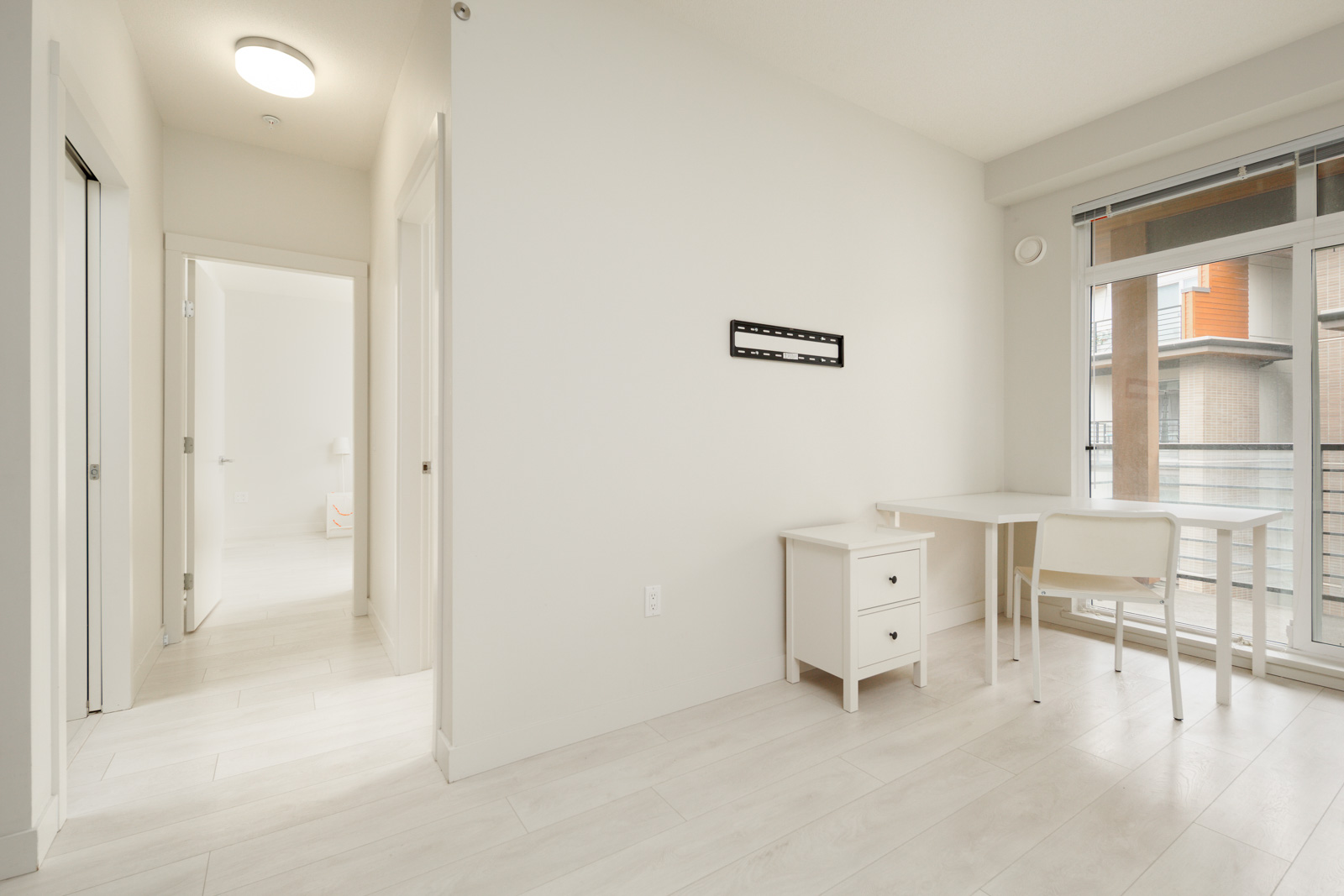Minimalist room with light wood flooring, a small white desk with a chair, and a window with blinds. An empty picture frame is on the wall. A hallway with doors is visible on the left.
