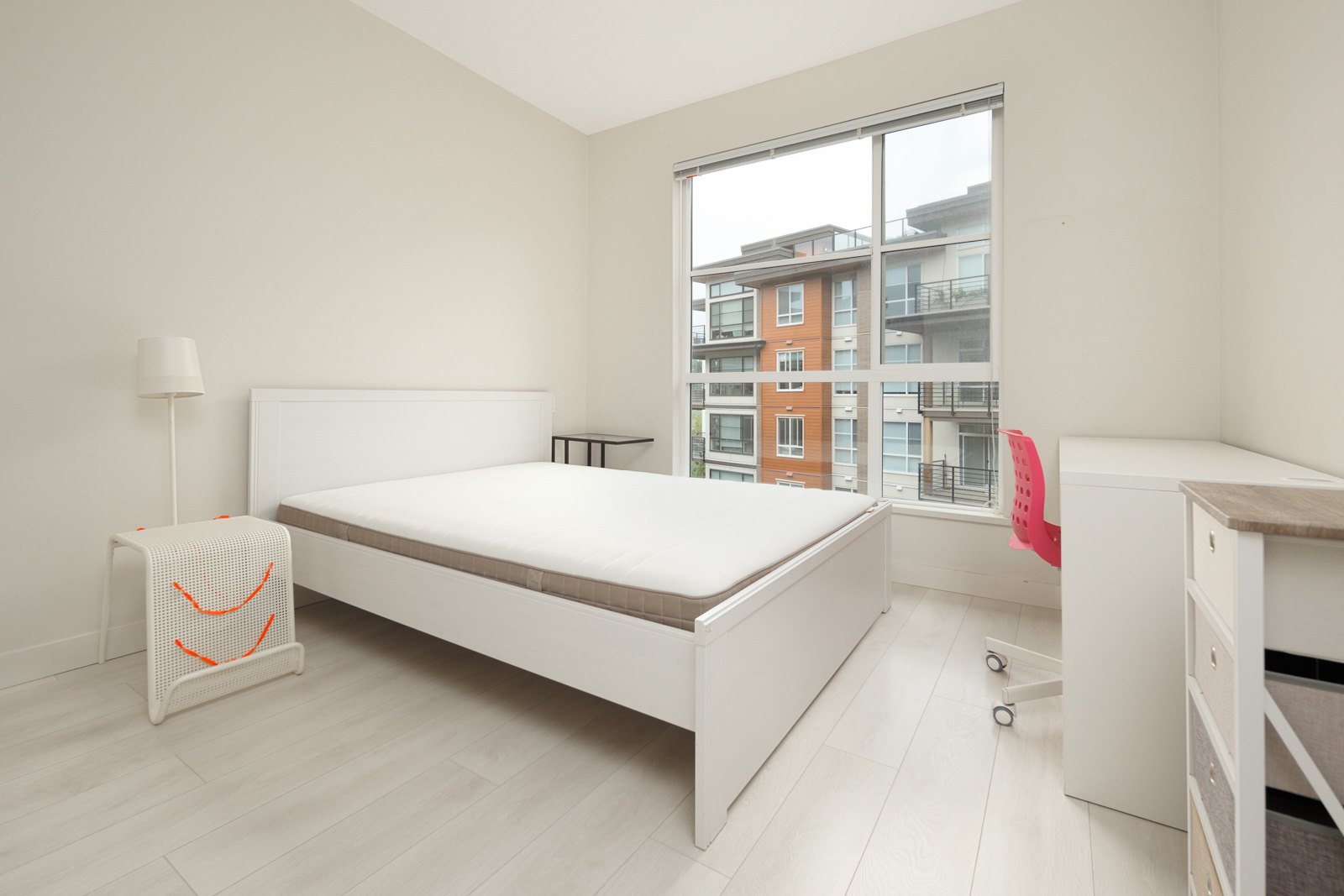 A minimalist bedroom with a white bed, white furniture, and a pink chair. A large window shows a view of apartment buildings outside.