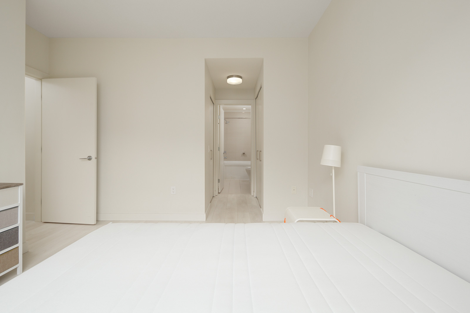 Minimalist bedroom with a white bed, nightstand, and lamp. Open door to a bathroom with a visible bathtub. Light walls and floor create a bright atmosphere.