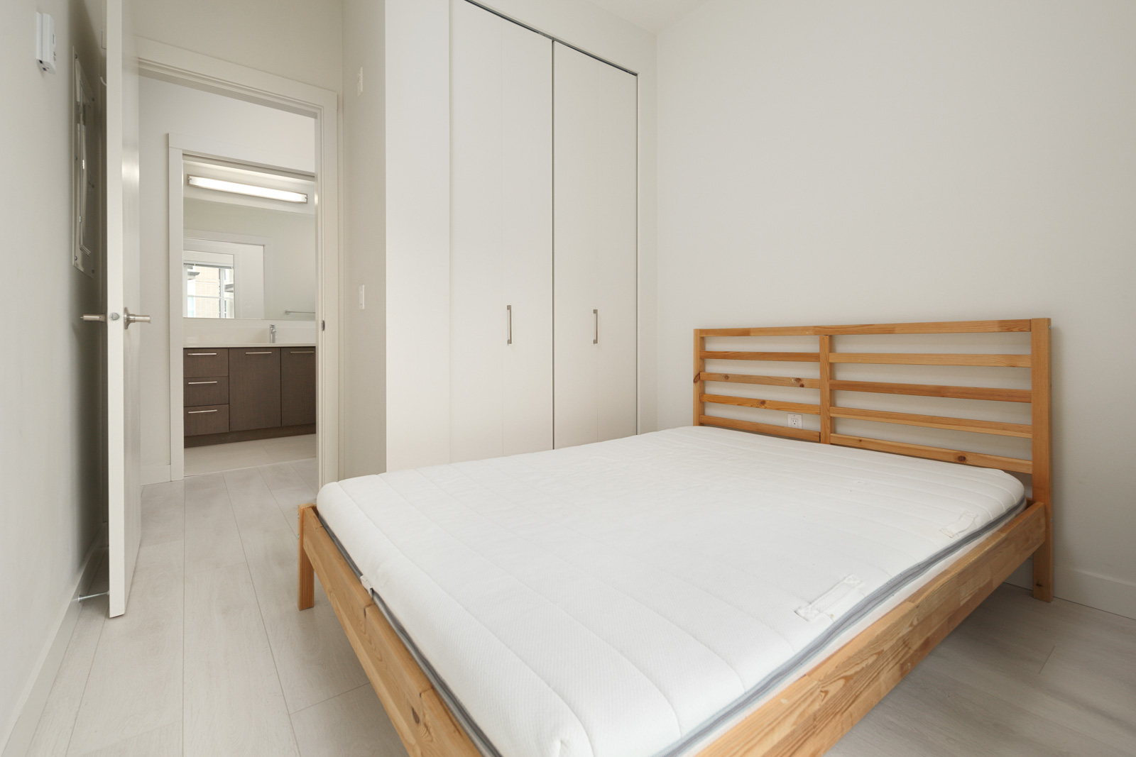 A minimalist bedroom with a wooden bed frame and white mattress. A closet with sliding doors is on the side, and a door leads to a bathroom with a vanity and mirror.