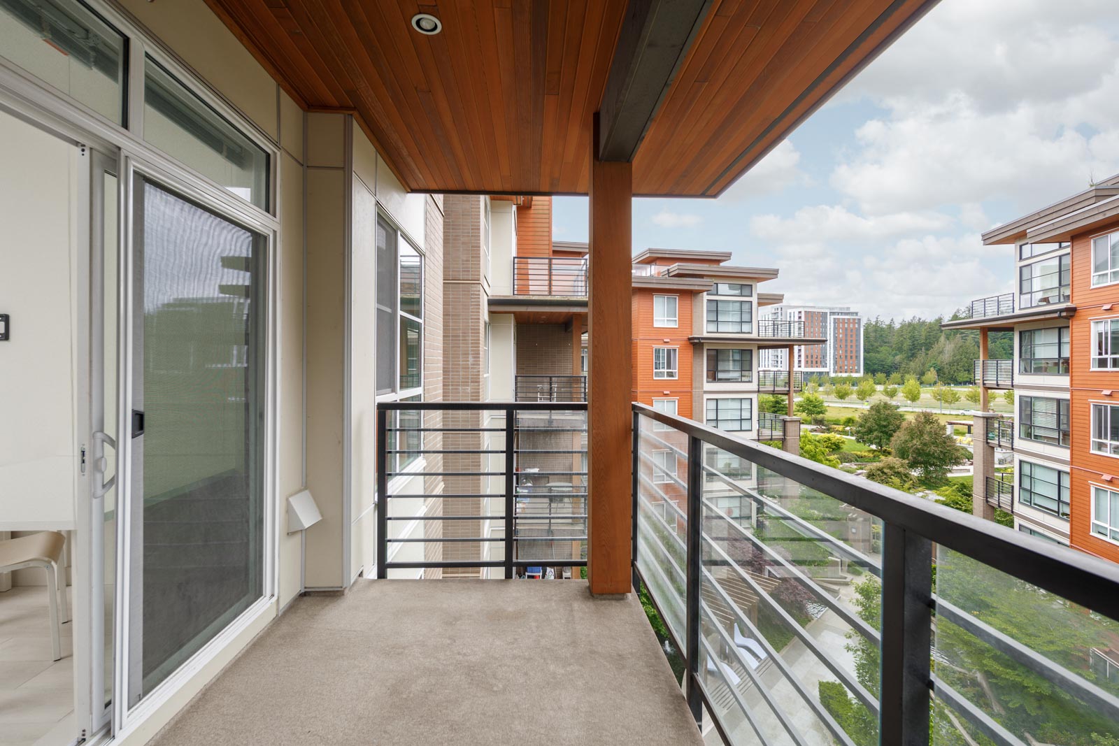 A modern apartment balcony with glass railing overlooks a landscaped courtyard and several multi-story buildings under a cloudy sky.