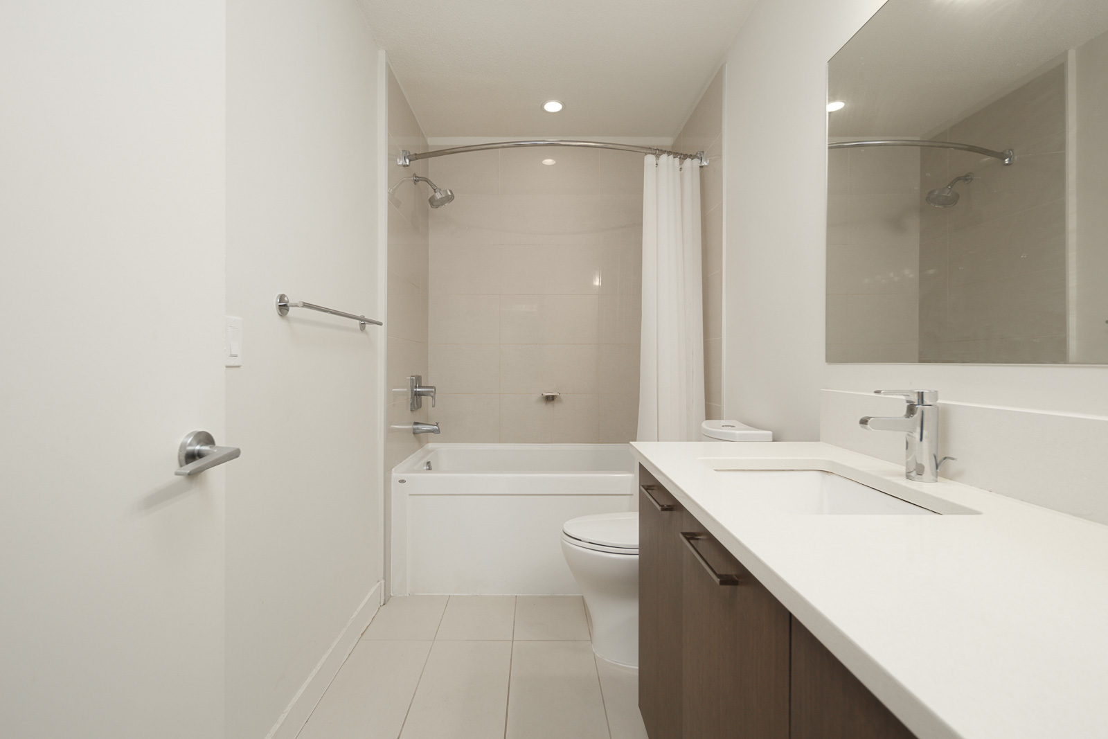 Modern bathroom with white walls and floor tiles, featuring a bathtub with a shower curtain, a toilet, a sink with a wide mirror, and wooden cabinetry.