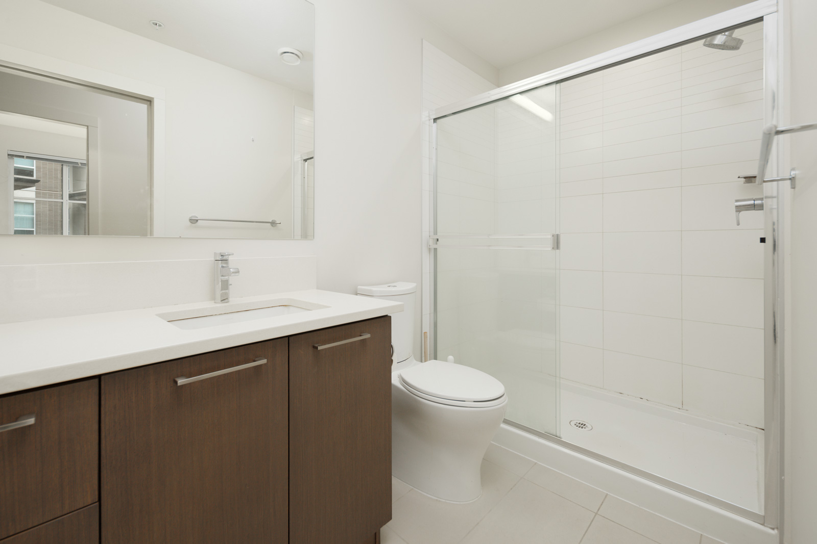 A modern bathroom with a wooden vanity, white countertop, sink, toilet, and a glass-enclosed shower.