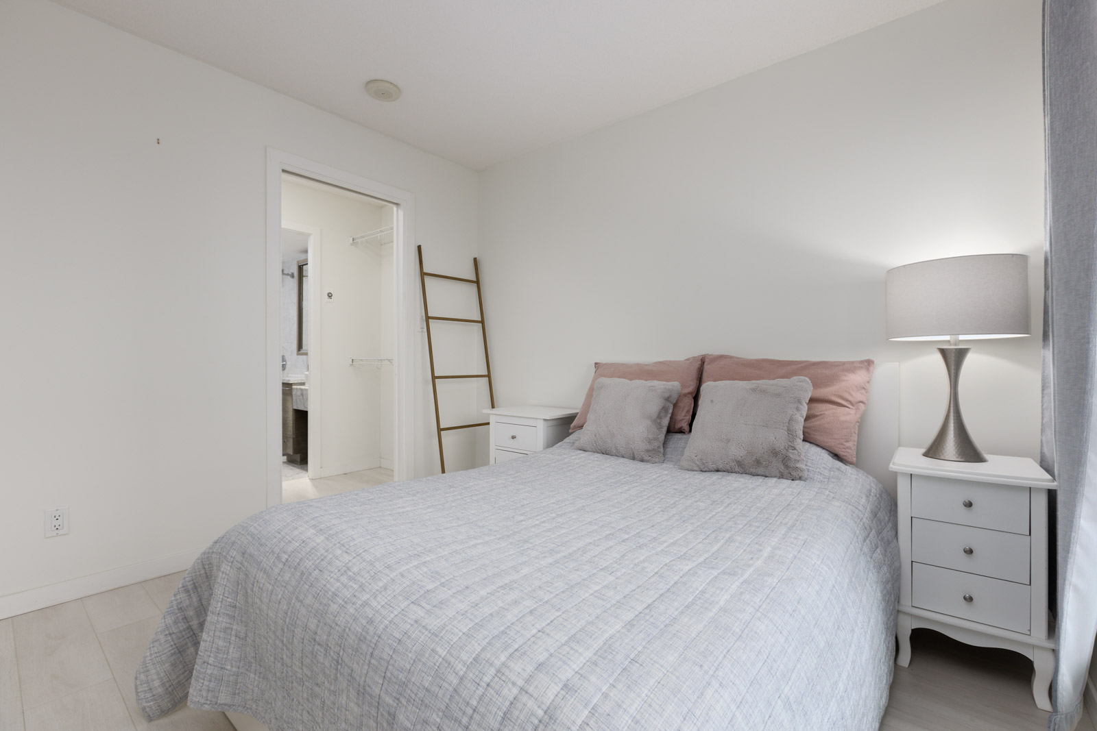 A neatly made bed with gray bedding and pink pillows is centered between two white nightstands; a lamp and ladder shelf are visible, with a bathroom through an open door.